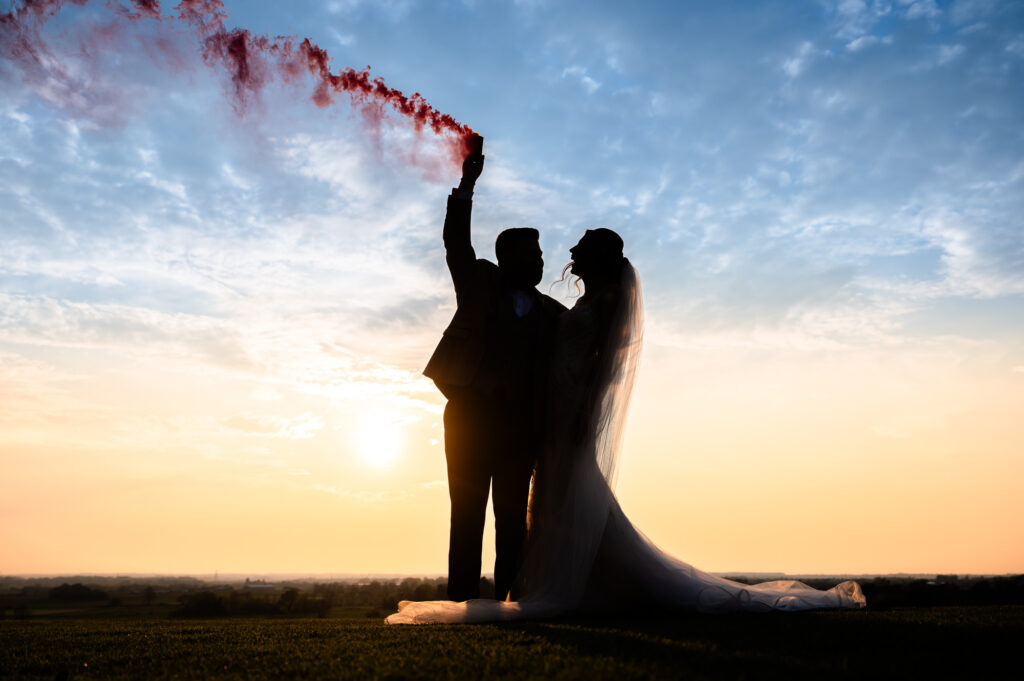 Groom holding an orange smoke grenade in the sunset with the bride next to him