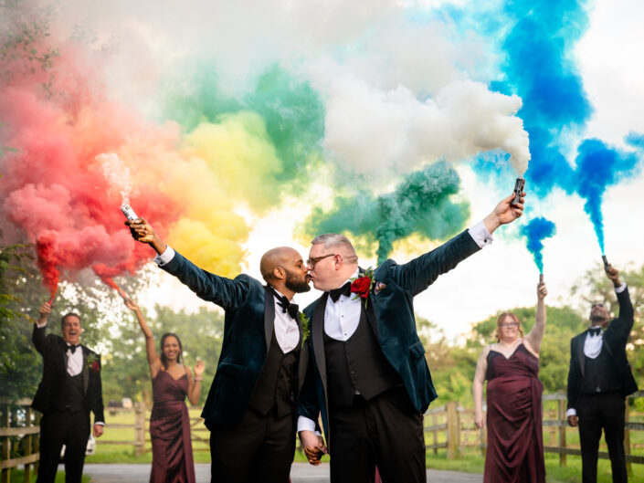 grooms kissing in front of rainbow coloured smoke flares at Owen House Wedding Barn