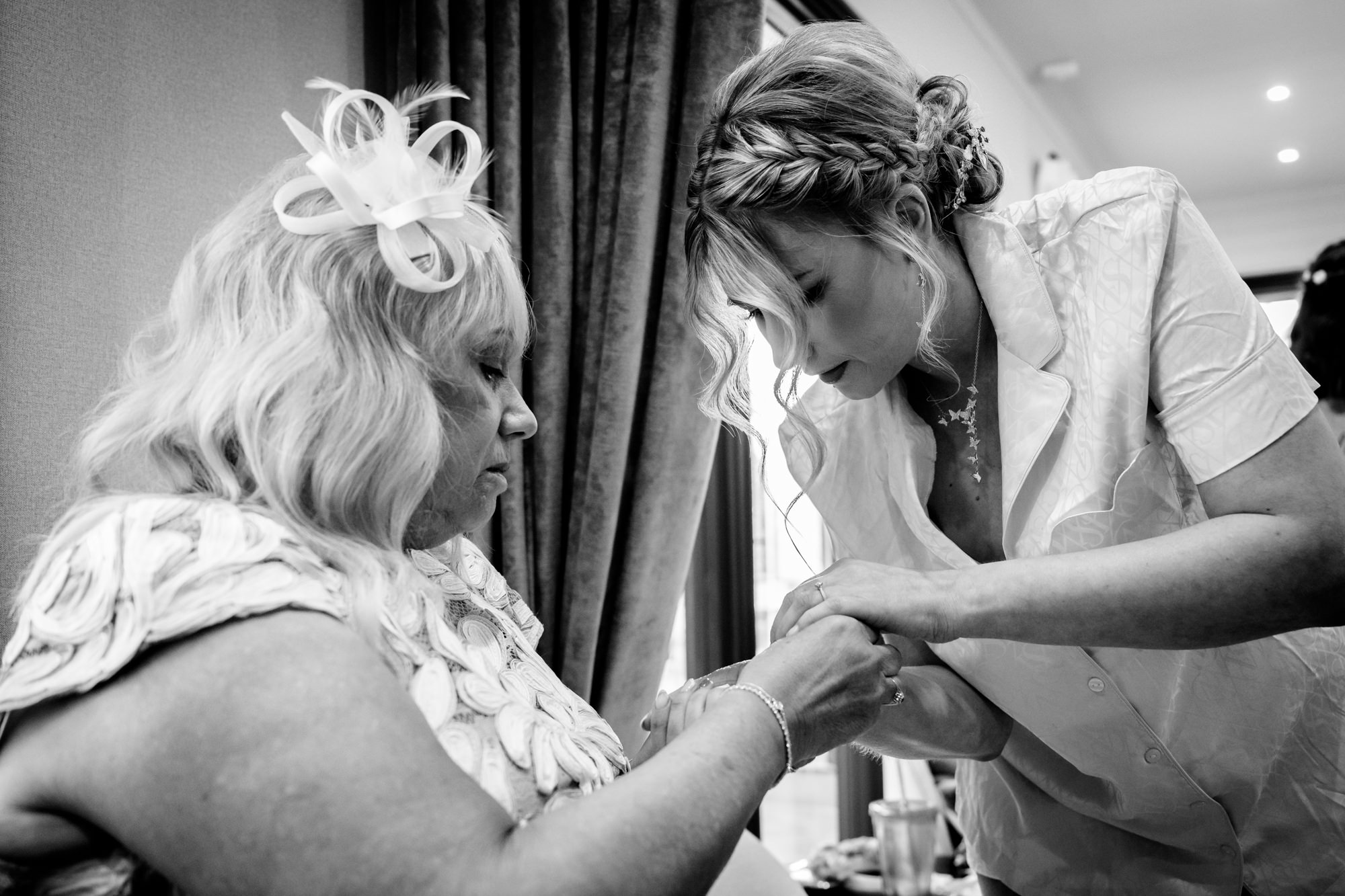 A black and white image of a bride in a white shirt helping an older woman fasten a bracelet on her wrist, during bridal prep in the prep pod at Moddershall Oaks.