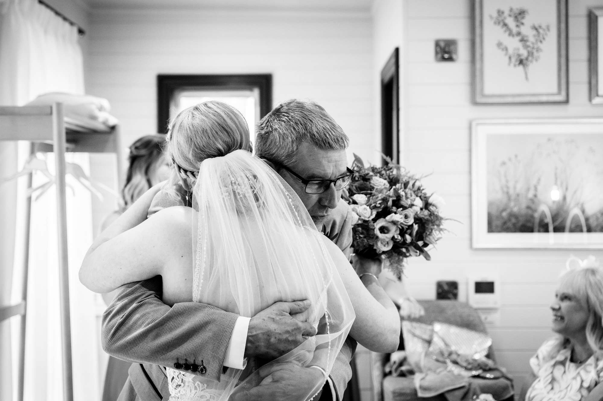 A black and white image of a the father of the bride hugging the bride in a wedding dress, holding a bouquet of flowers, during bridal prep in the prep pod at Moddershall Oaks.