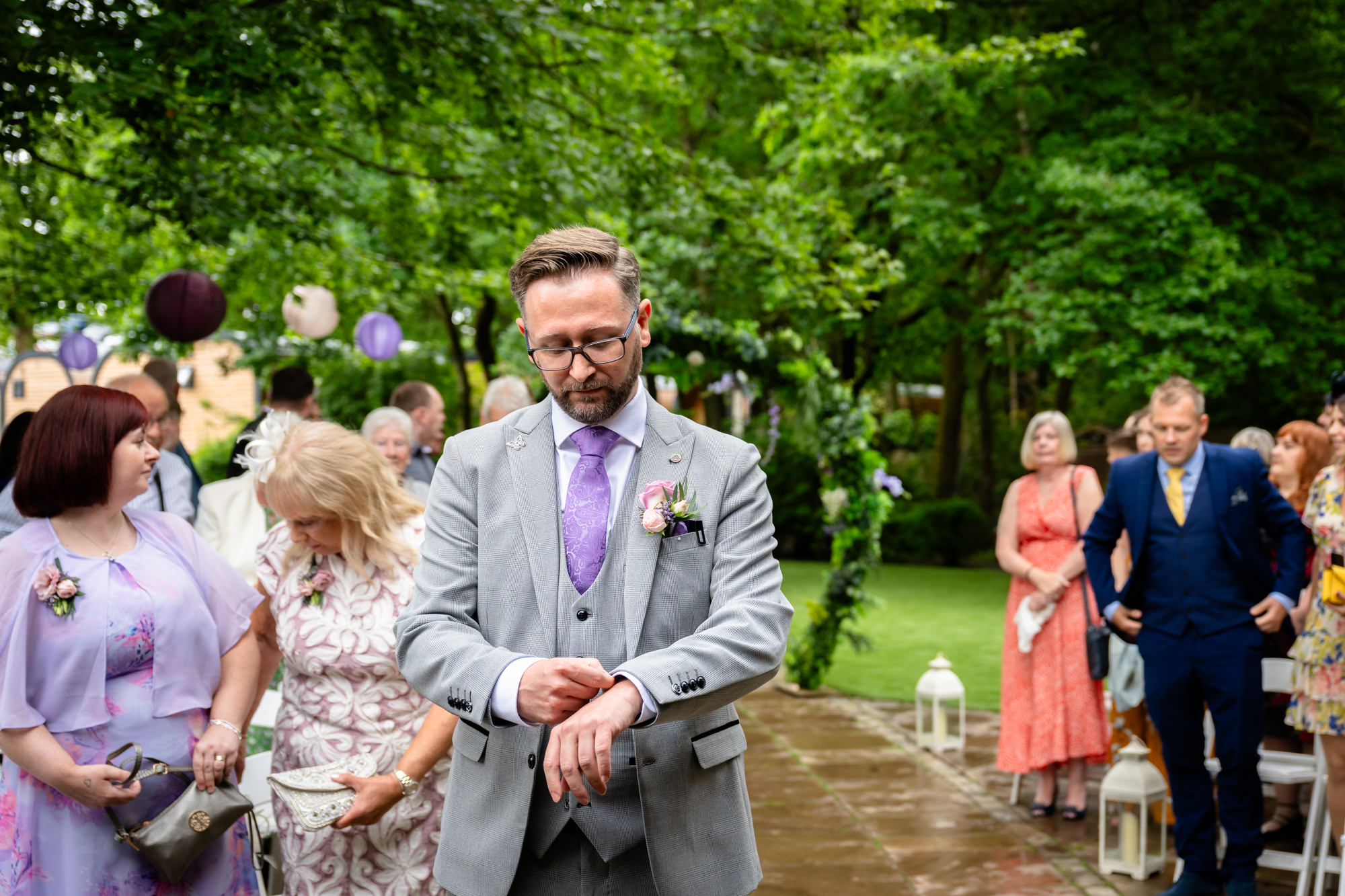 A groom in a grey suit and purple tie walks outdoors at Moddershall Oaks, looking down at his wrist, surrounded by guests during the outdoor ceremony.