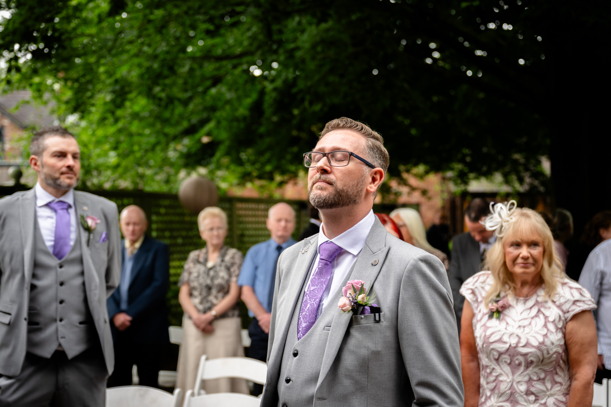 A groom in a grey suit and purple tie looks upwards, with guests and greenery in the background, awaiting the bride at the outdoor ceremony at Moddershall Oaks.