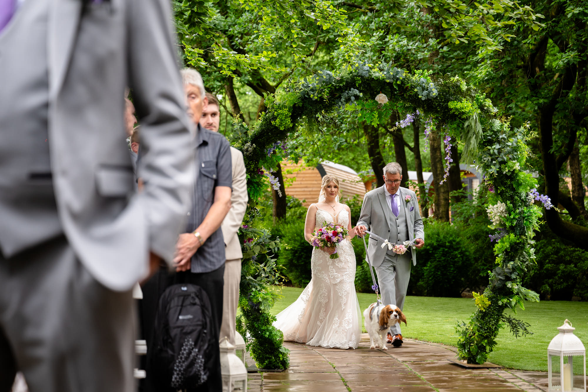 A bride in a white gown walks down a green aisle with her father and dog, towards the wedding arch at Moddershall Oaks, carrying a bouquet during the outdoor ceremony.