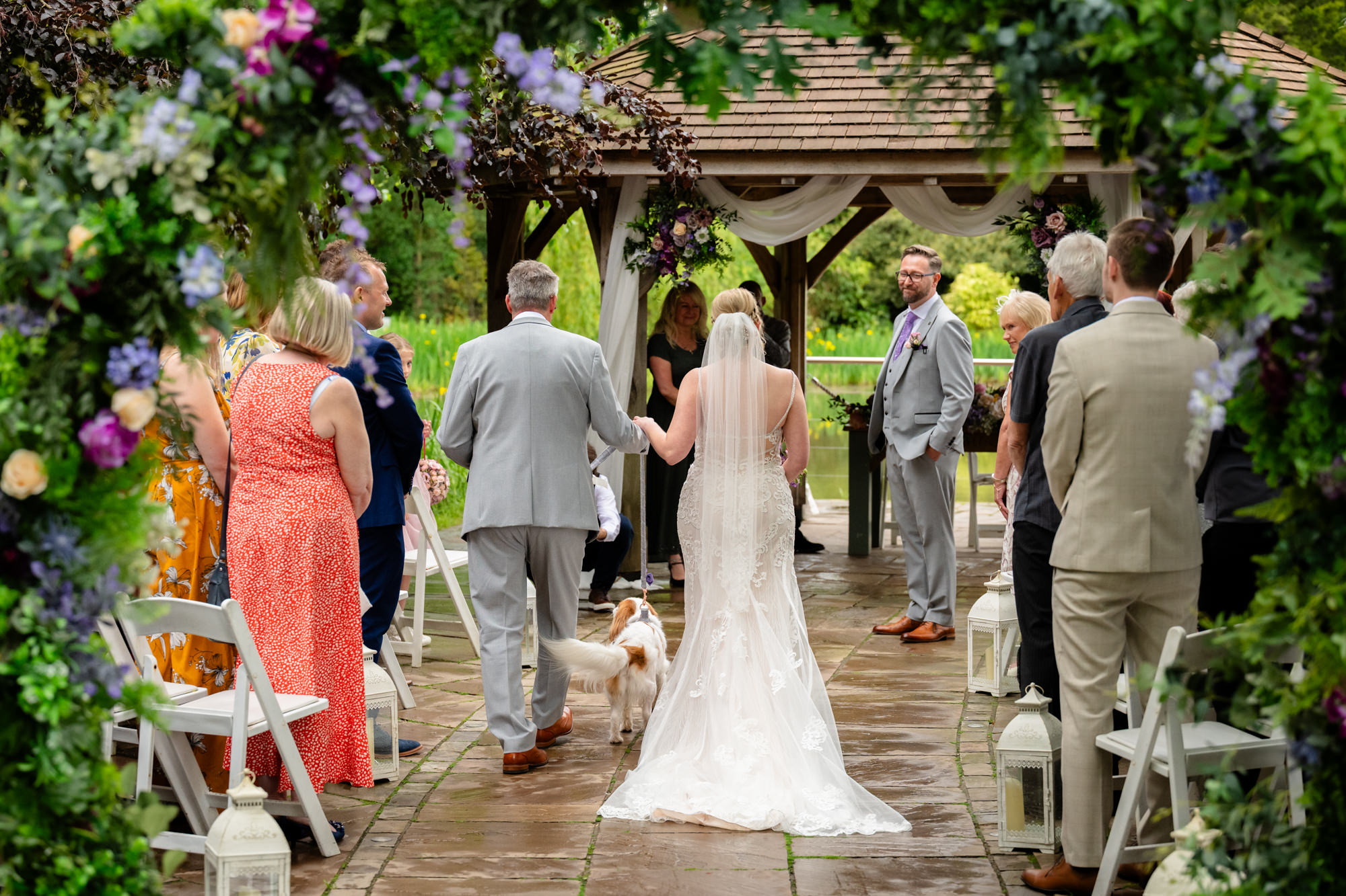 The bride, viewed from behind, walking down the aisle towards a decorated gazebo at Moddershall Oaks, with guests seated on both sides during the outdoor ceremony.