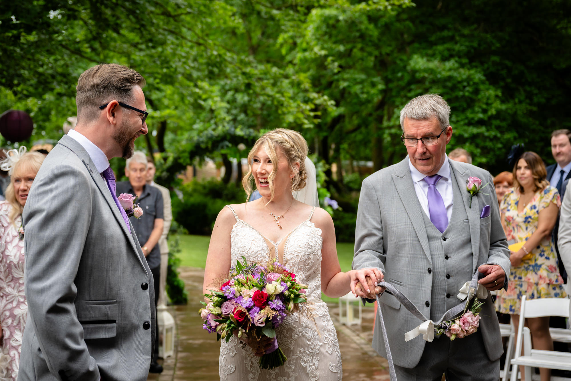 The bride, looking happy, walks down the aisle arm in arm with her father, while the groom awaits at the outdoor ceremony at Moddershall Oaks.
