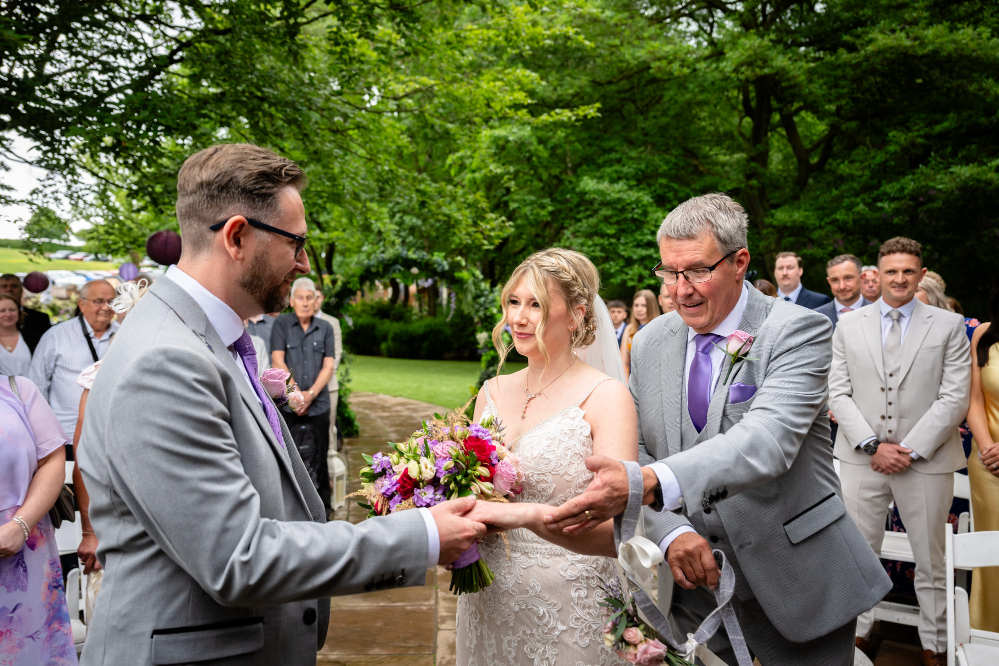 A smiling bride is handed over by her father to the waiting groom at the end of the aisle, during the outdoor ceremony at Moddershall Oaks.