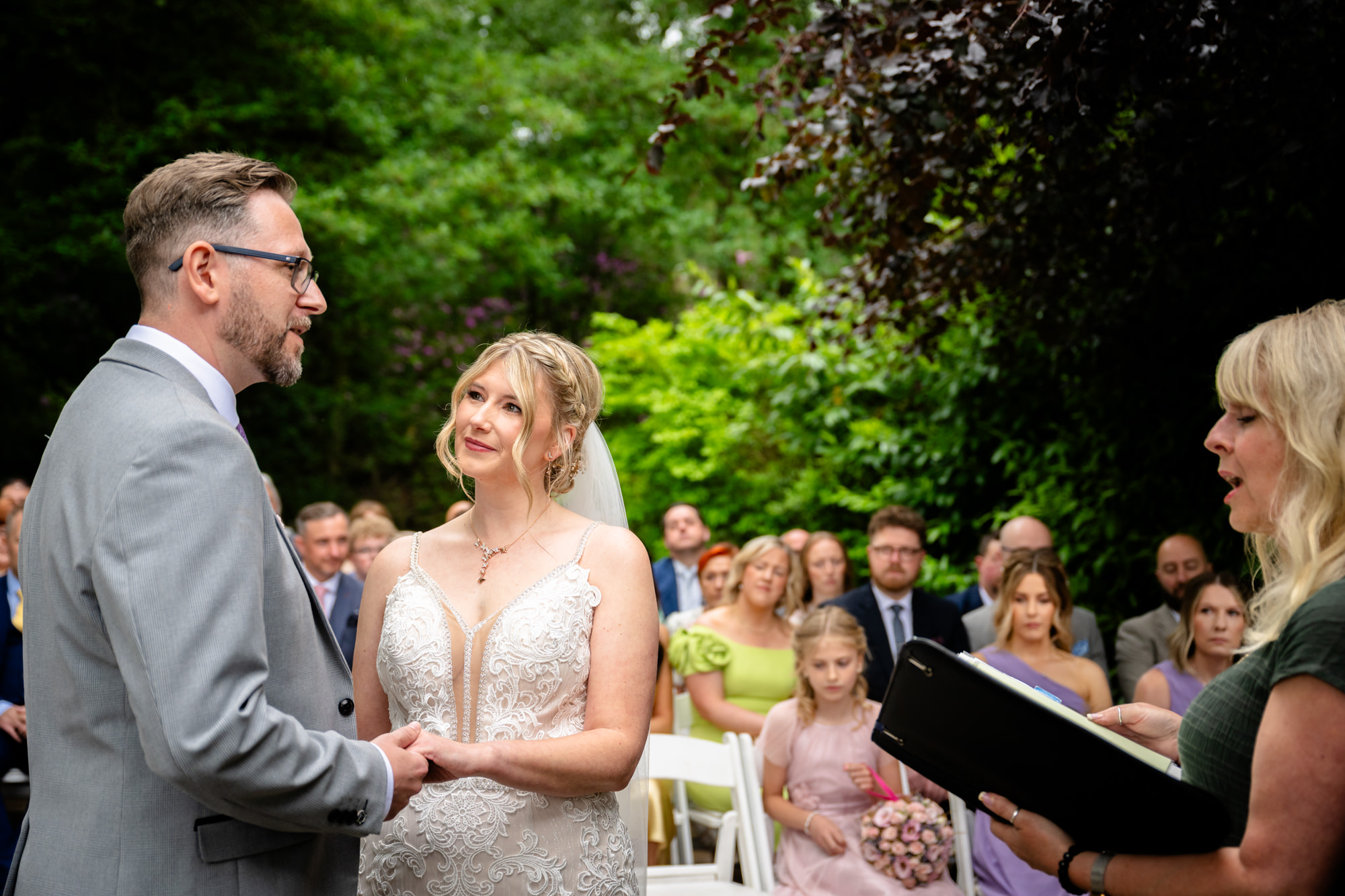 The bride and groom stand facing each other under the Oak Pagoda at Moddershall Oaks, with a registrar to the right and guests in the background during the outdoor ceremony.