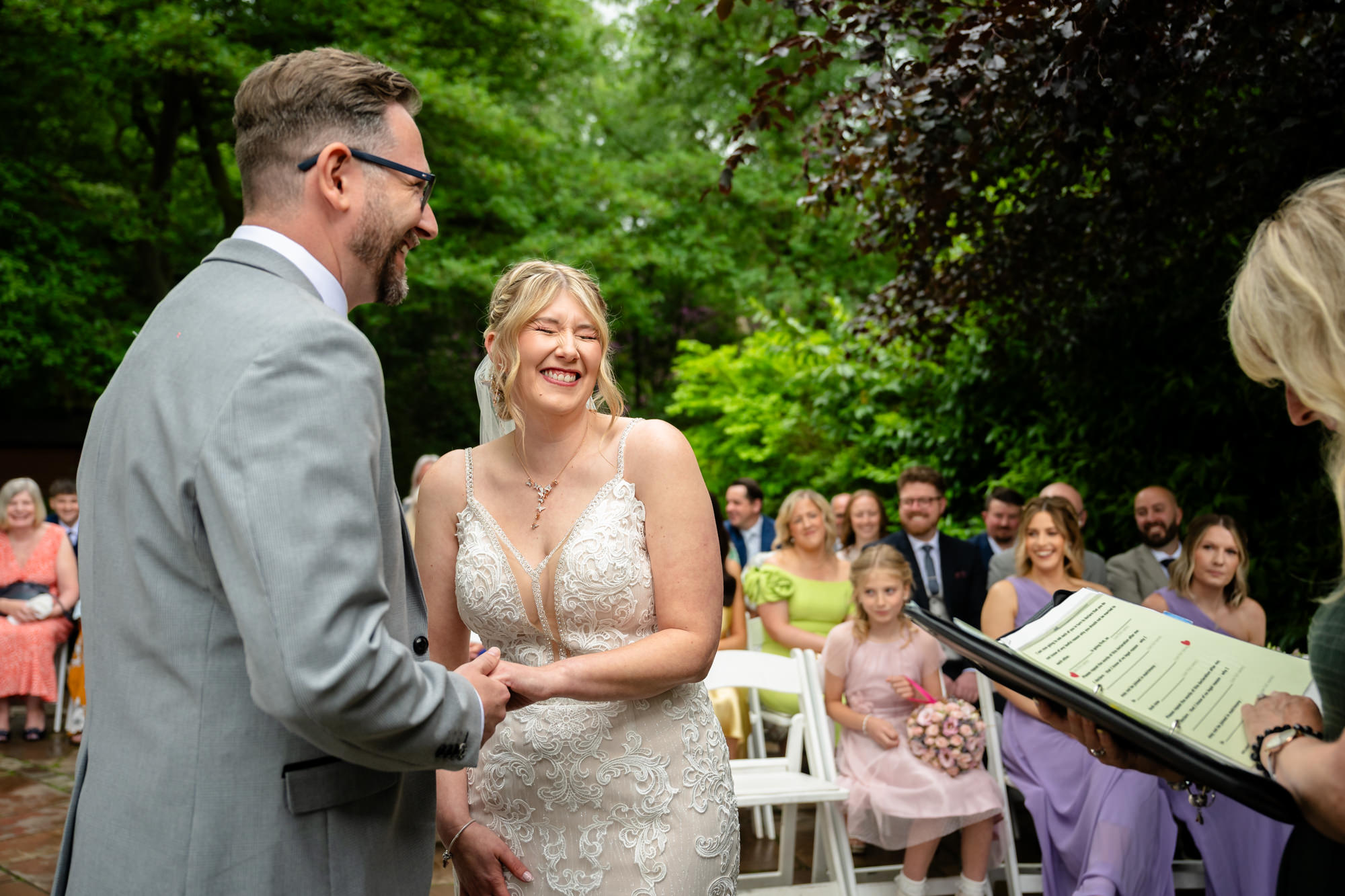 The bride and groom stand smiling at each other during their outdoor ceremony at Moddershall Oaks, surrounded by greenery and guests.