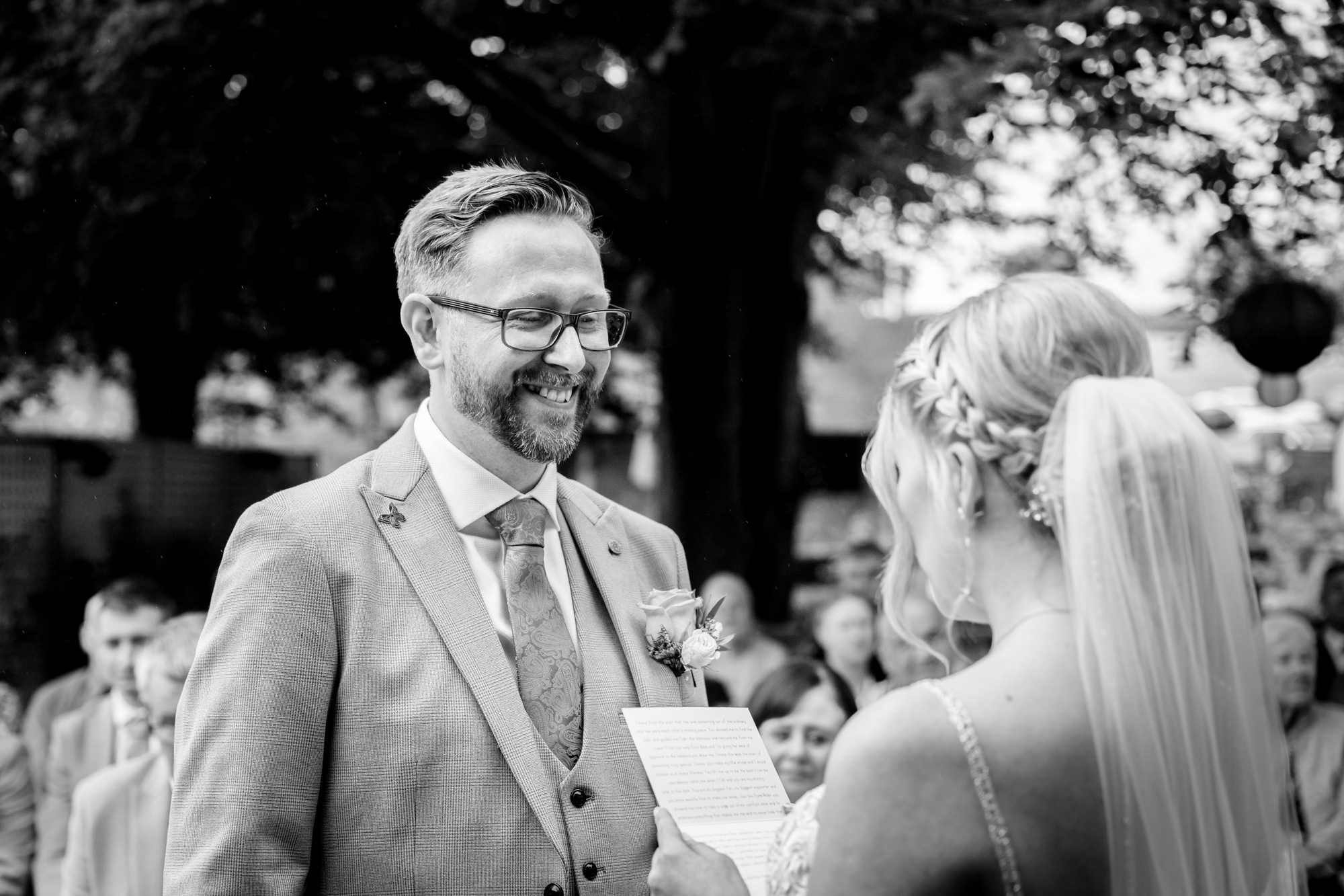 A black and white image of the groom smiling at the bride during the outdoor wedding ceremony at Moddershall Oaks.