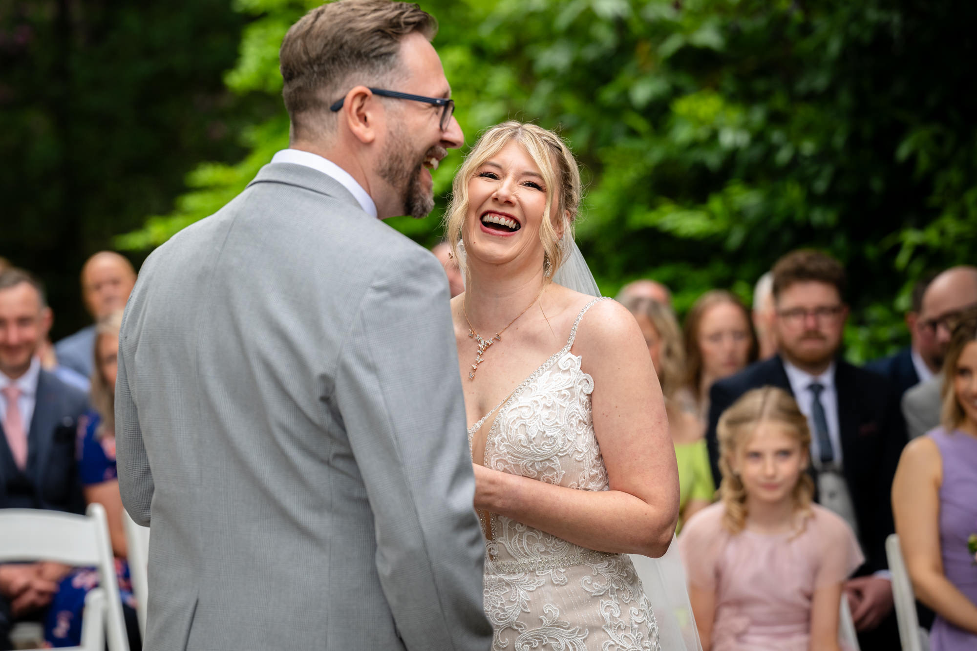 The bride and groom share a joyful laugh during their wedding ceremony at Moddershall Oaks, with guests blurred in the background.