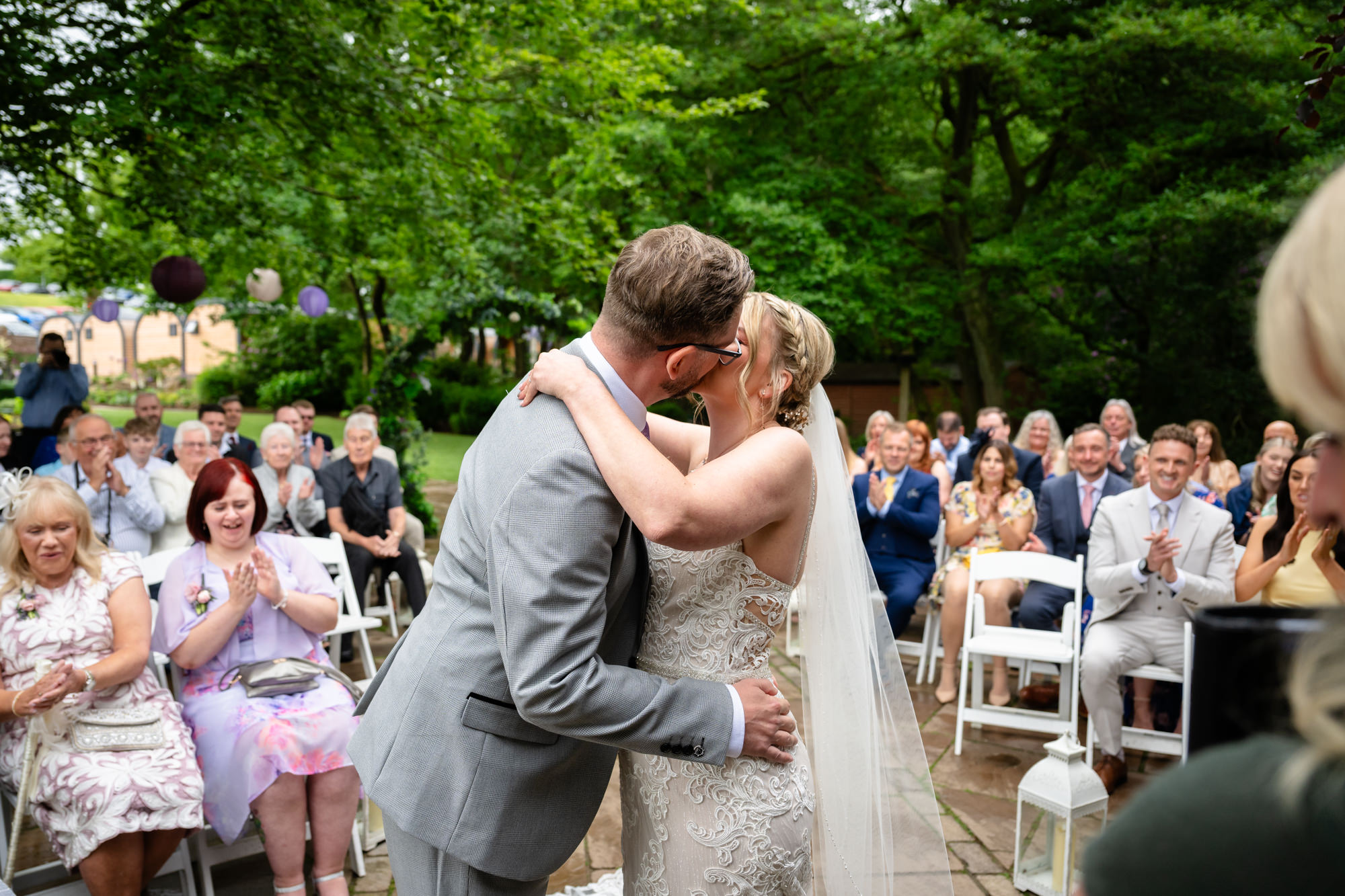 The bride and groom share their first kiss as a married couple, surrounded by applauding guests outdoors at Moddershall Oaks.