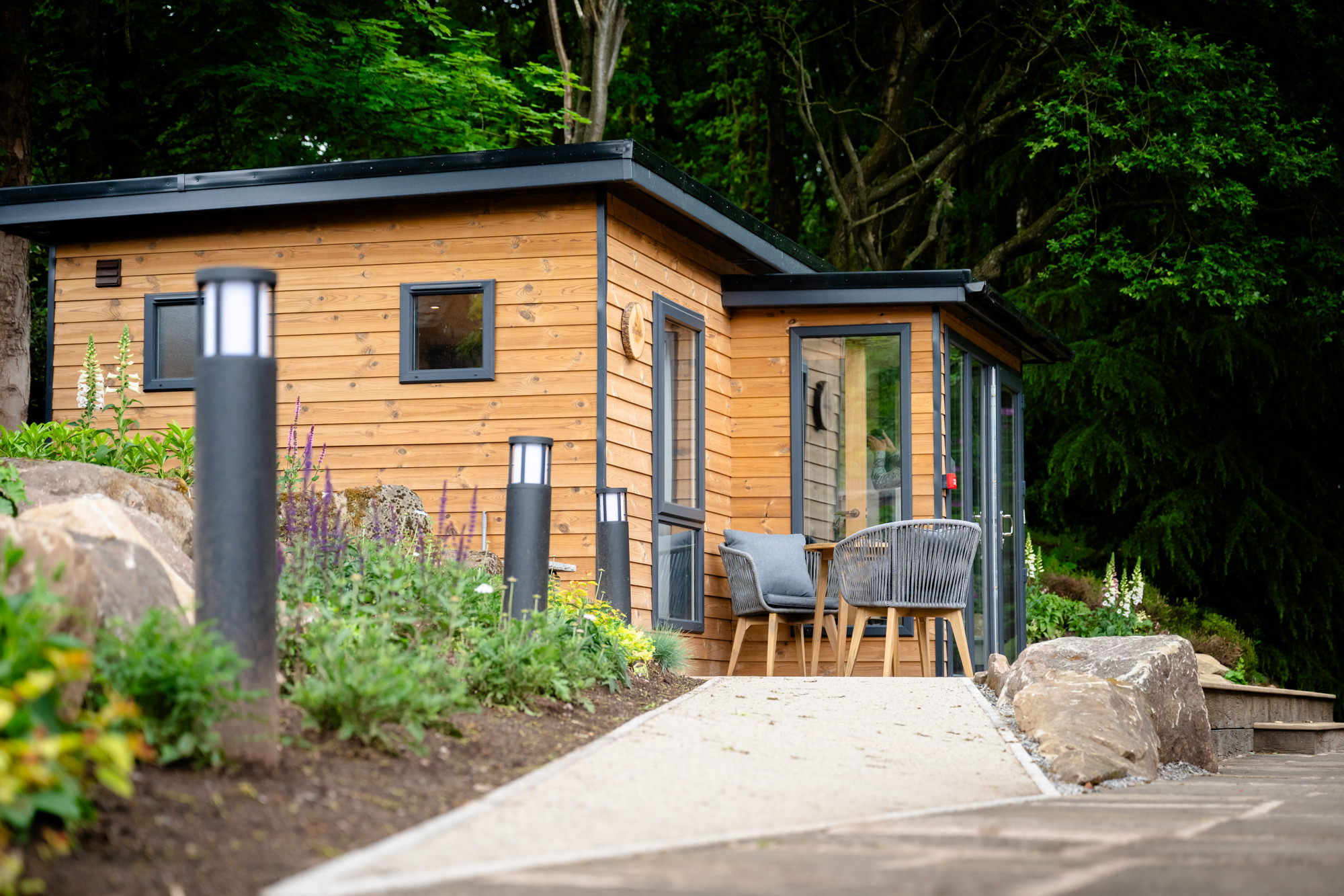 A modern wooden cabin with large windows and outdoor seating, surrounded by lush green foliage and stone landscaping, part of the bridal prep pod at Moddershall Oaks.