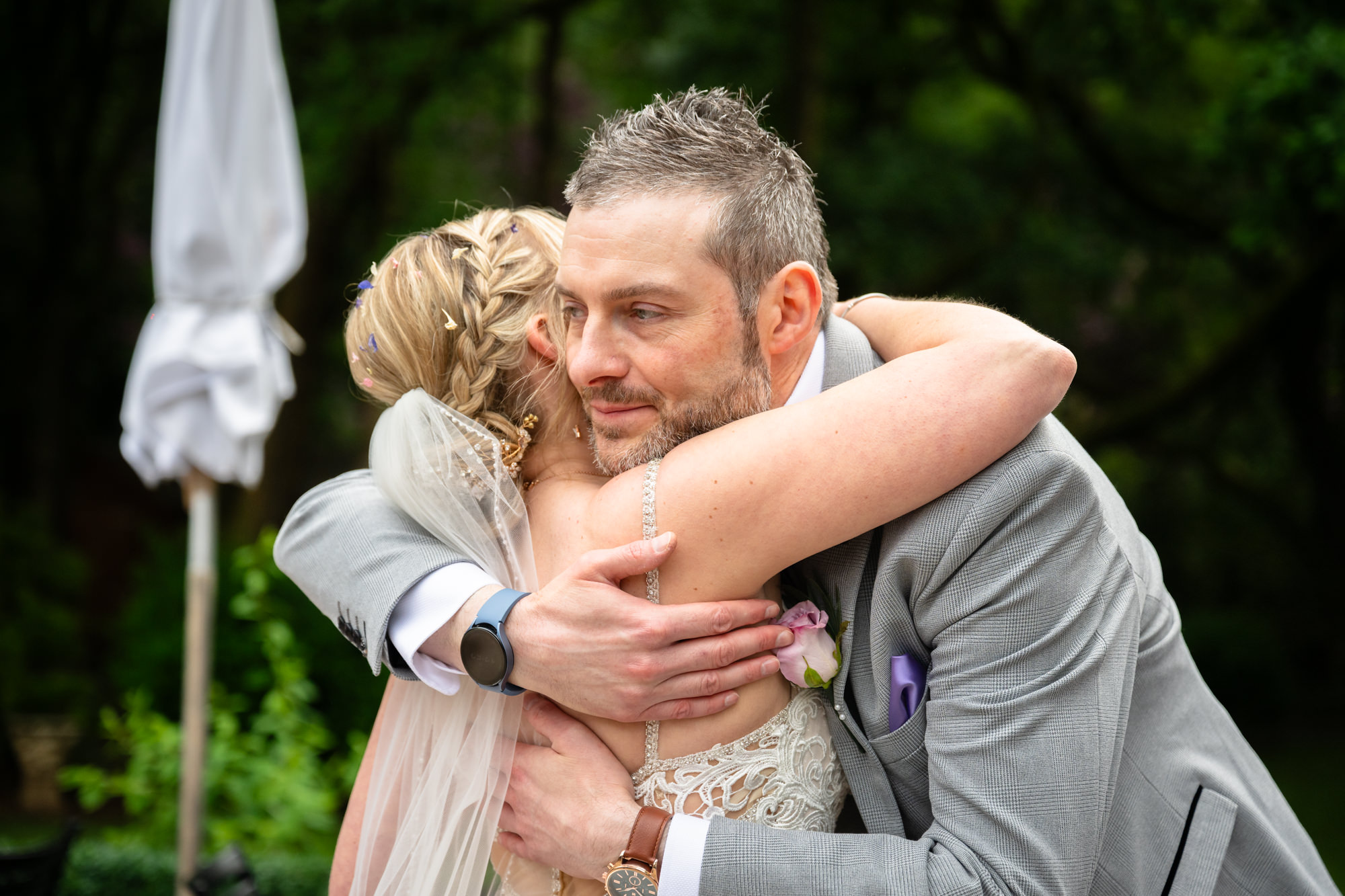 A close-up of a groomsman embracing the bride warmly, both smiling, surrounded by a green outdoor setting at Moddershall Oaks.