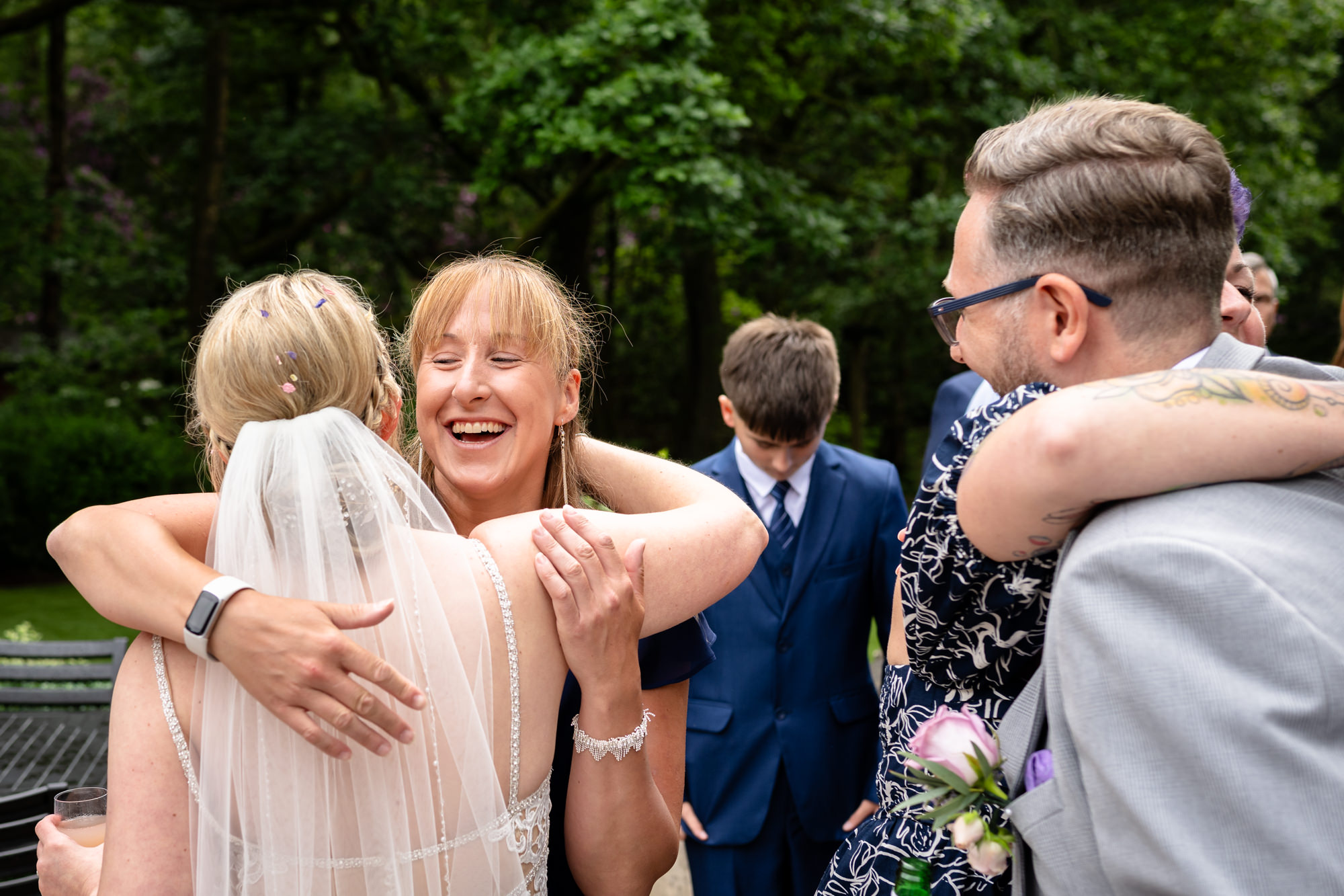Two women embrace at Moddershall Oaks, with one laughing joyfully, against a backdrop of greenery and other guests.