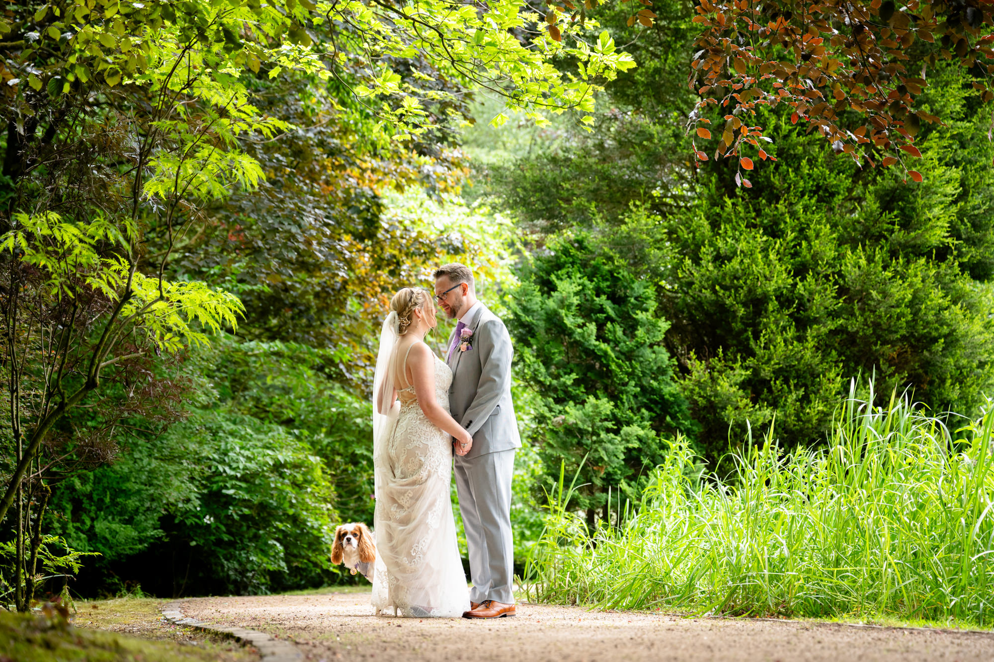The bride and groom stand facing each other under a canopy of green trees, holding hands, in a romantic relaxed portrait photo around the lake at Moddershall Oaks.