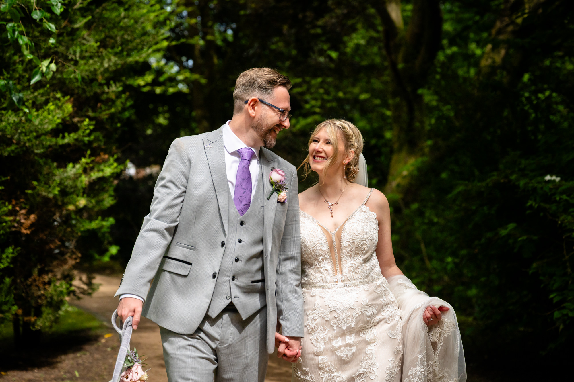 The bride and groom walk hand in hand down a path, looking at each other and smiling, surrounded by green trees, during relaxed portrait photos around the lake at Moddershall Oaks.