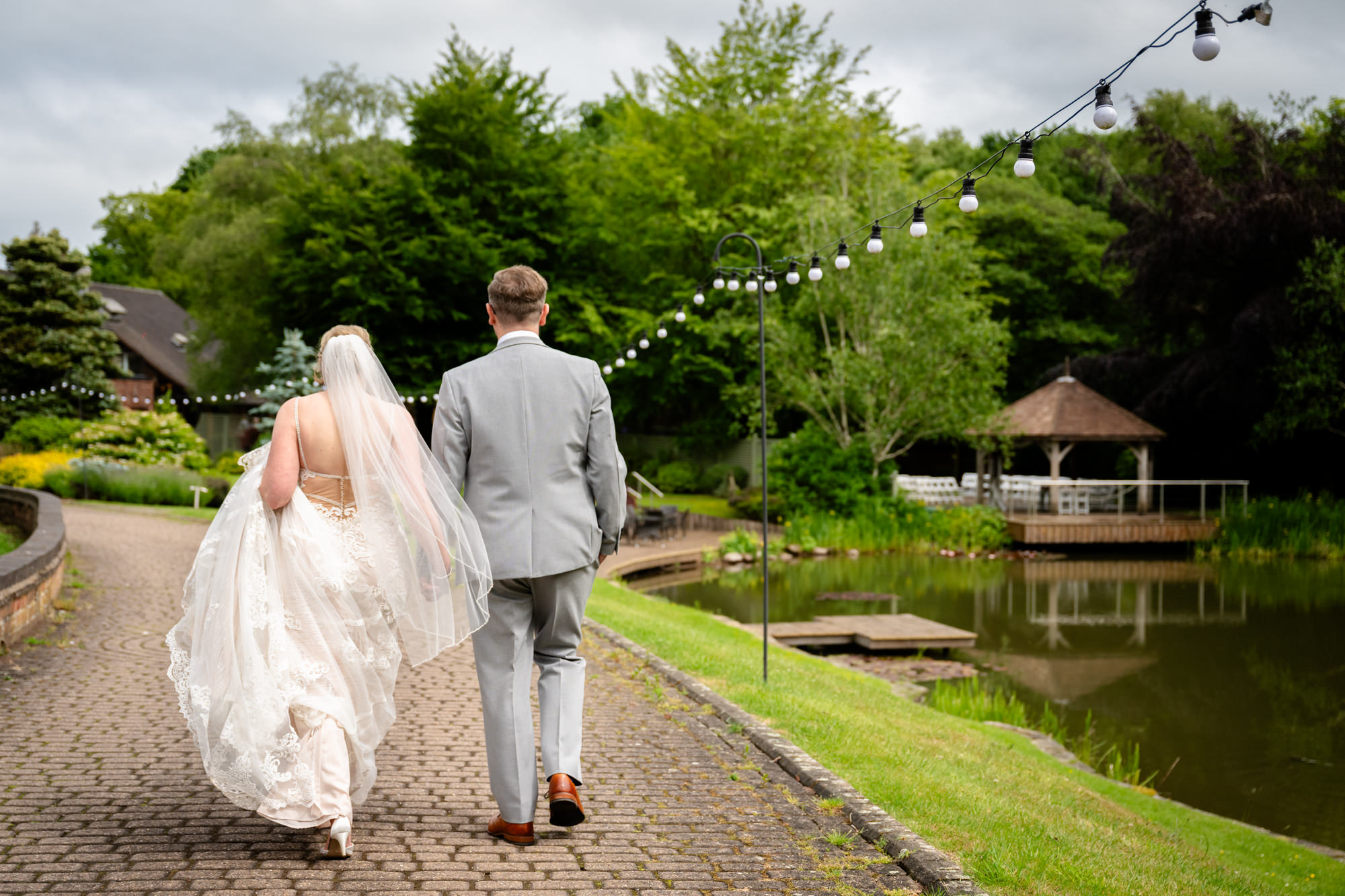 The bride and groom, viewed from behind, walk away from the camera on a paved path, with a pond and distant building in the background, during relaxed portrait photos around the lake at Moddershall Oaks.