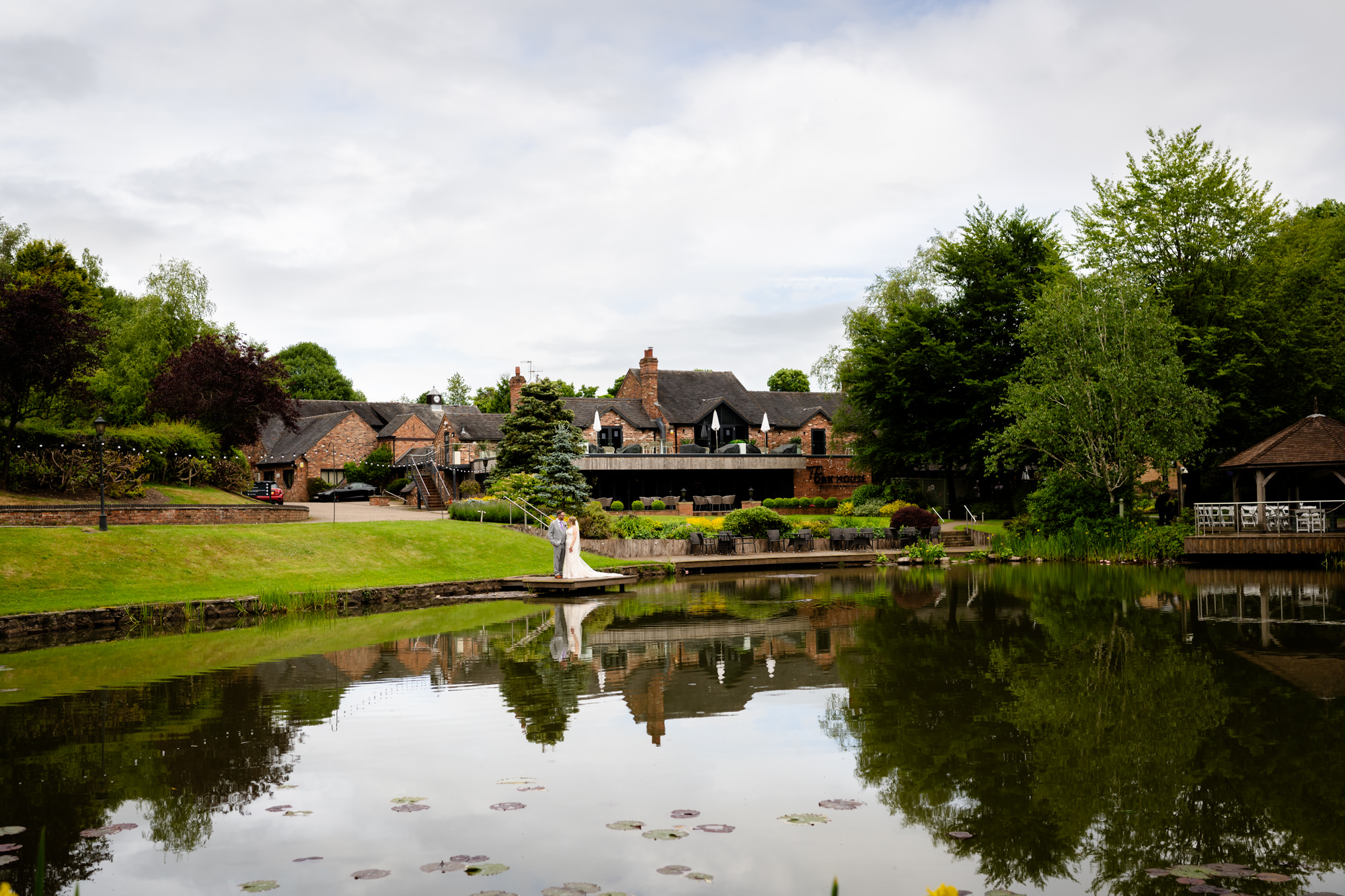 A wide shot of Moddershall Oaks, featuring a tranquil pond reflecting the surrounding green trees and the building in the distance, during relaxed portrait photos around the lake.