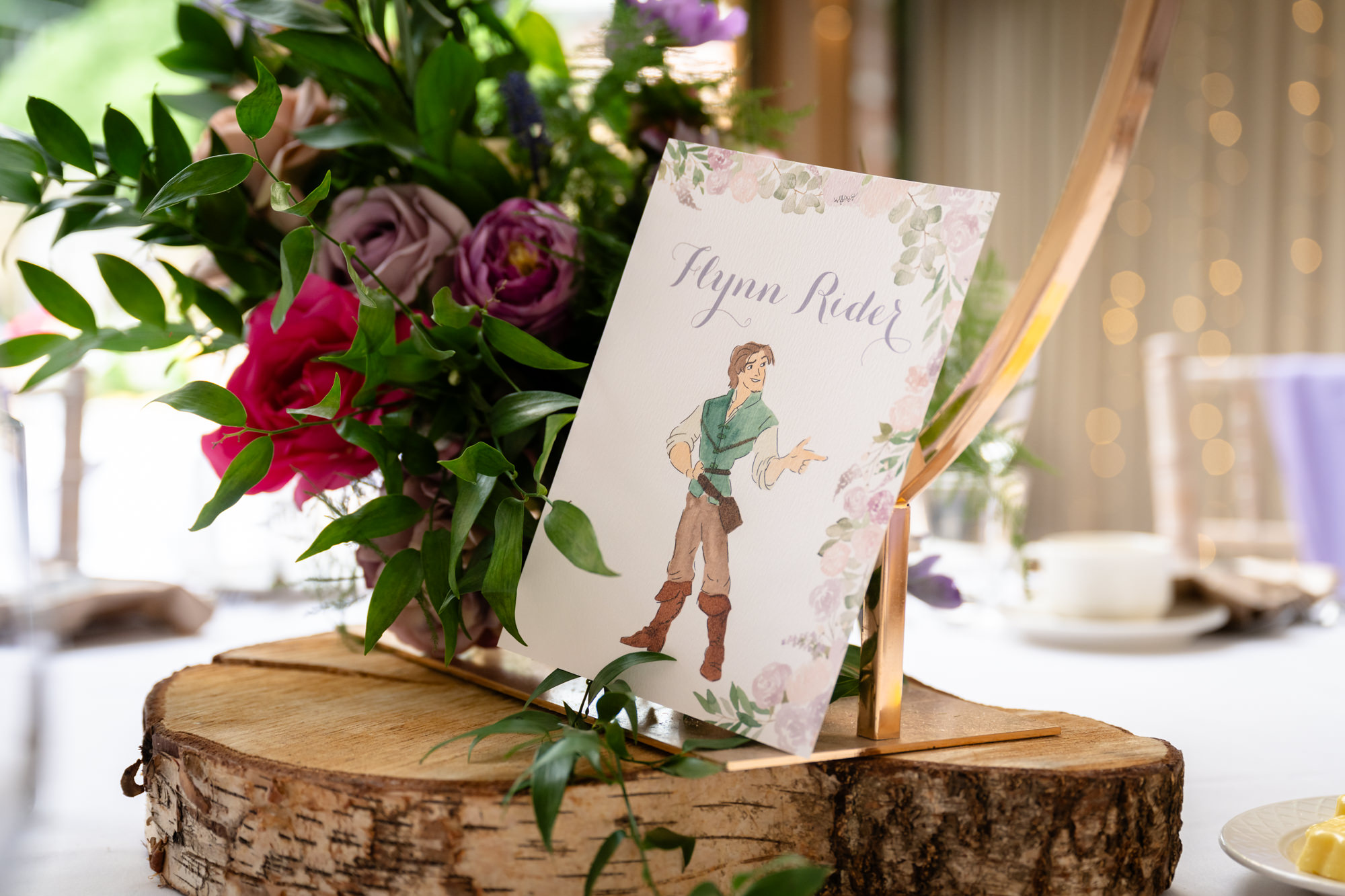 A wedding table centerpiece at Moddershall Oaks featuring a slice of wood, green foliage, red flowers, and an illustrated place card of a couple, reflecting Disney's Tangled-themed wedding details.
