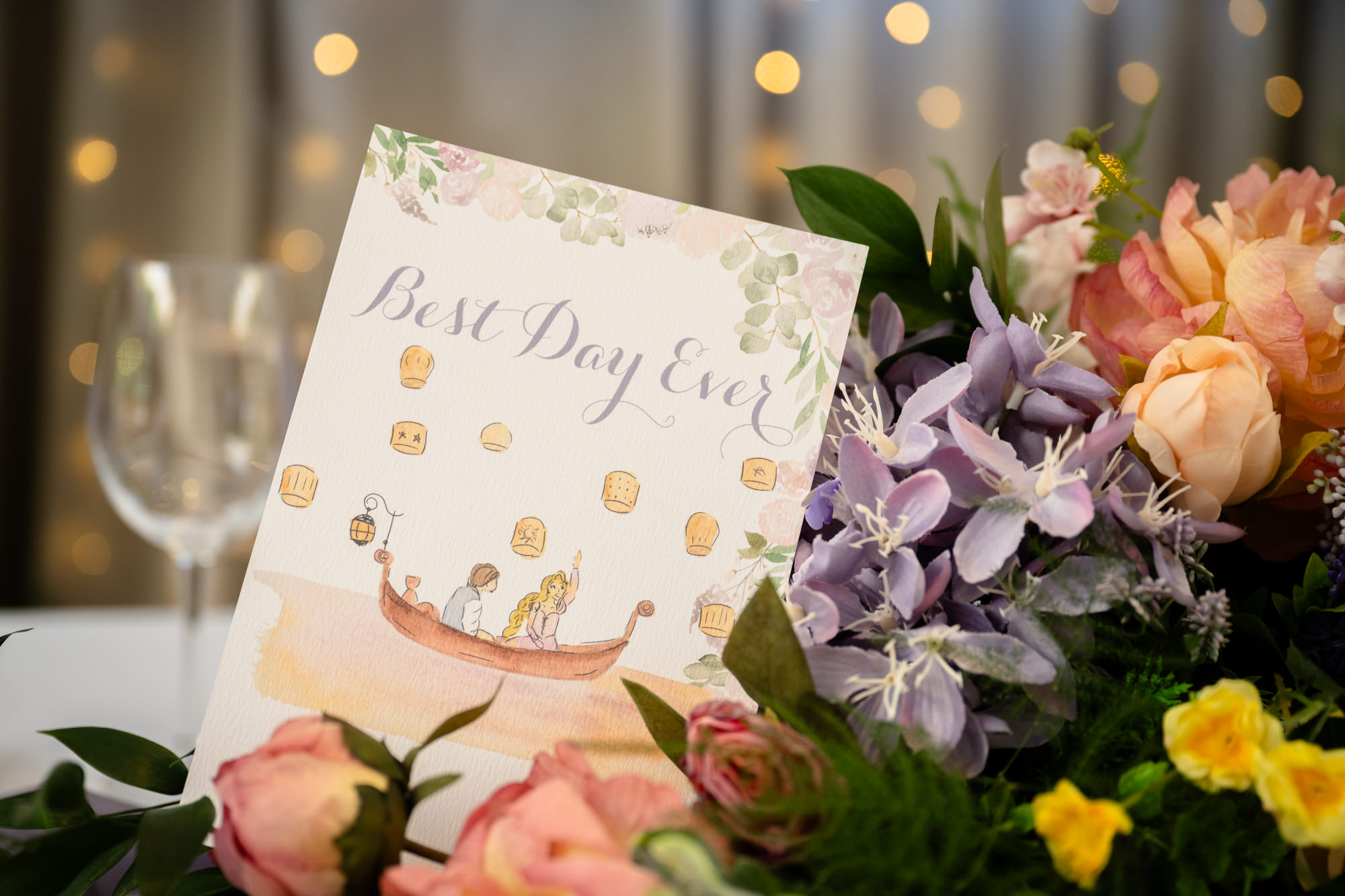 A close-up of a wedding table setting at Moddershall Oaks with a white menu card, surrounded by green leaves and pink and orange floral arrangements, showcasing Disney's Tangled-themed wedding details.