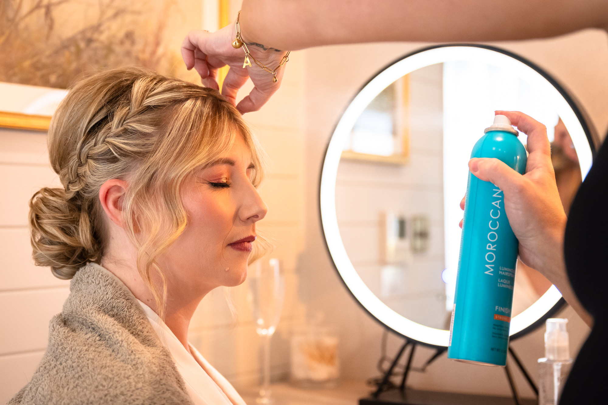 A close-up of a bride with intricate braided hair receiving a final spray of hairspray in front of an illuminated mirror, in the prep pod at Moddershall Oaks.