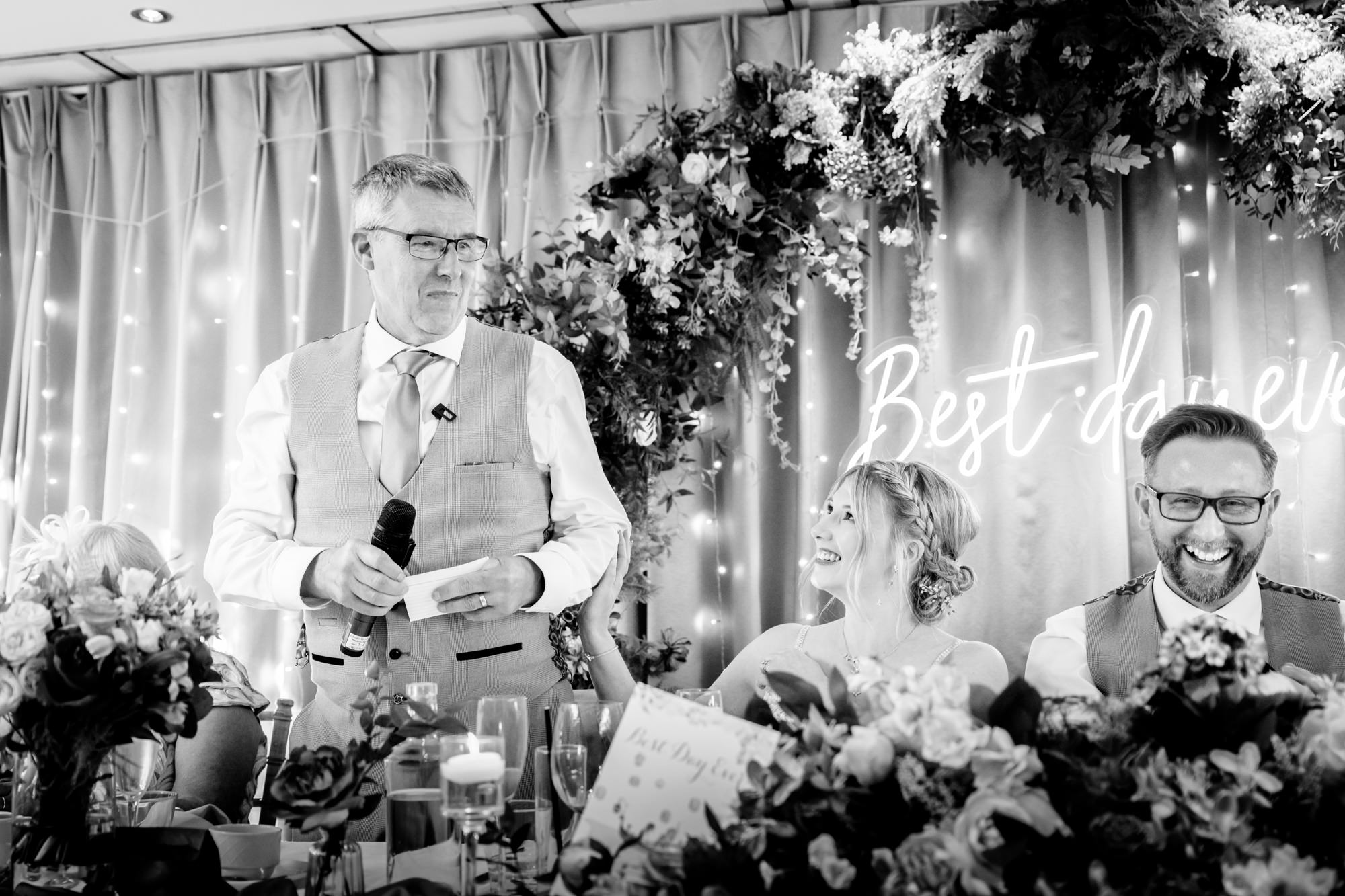 A black and white image of the wedding party seated at the head table at Moddershall Oaks, with a floral archway behind them, during the speeches.