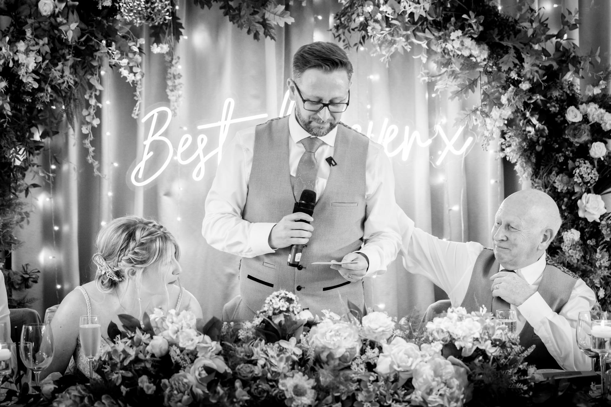 A black and white image of the groom speaking into a microphone, with the bridal party seated at the head table behind flowers, during speeches at Moddershall Oaks.