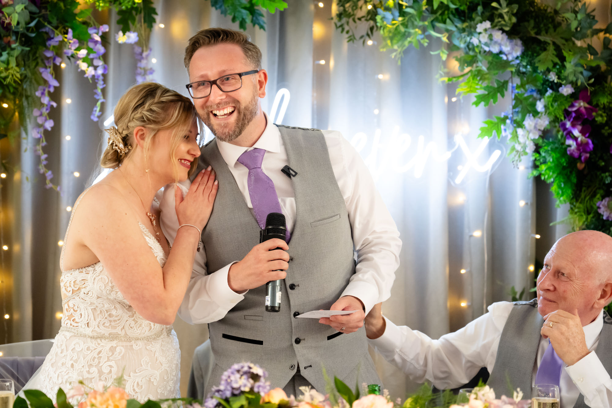 The bride playfully hides her face while the groom laughs and speaks into a microphone during his speech, surrounded by flowers and greenery, at Moddershall Oaks.