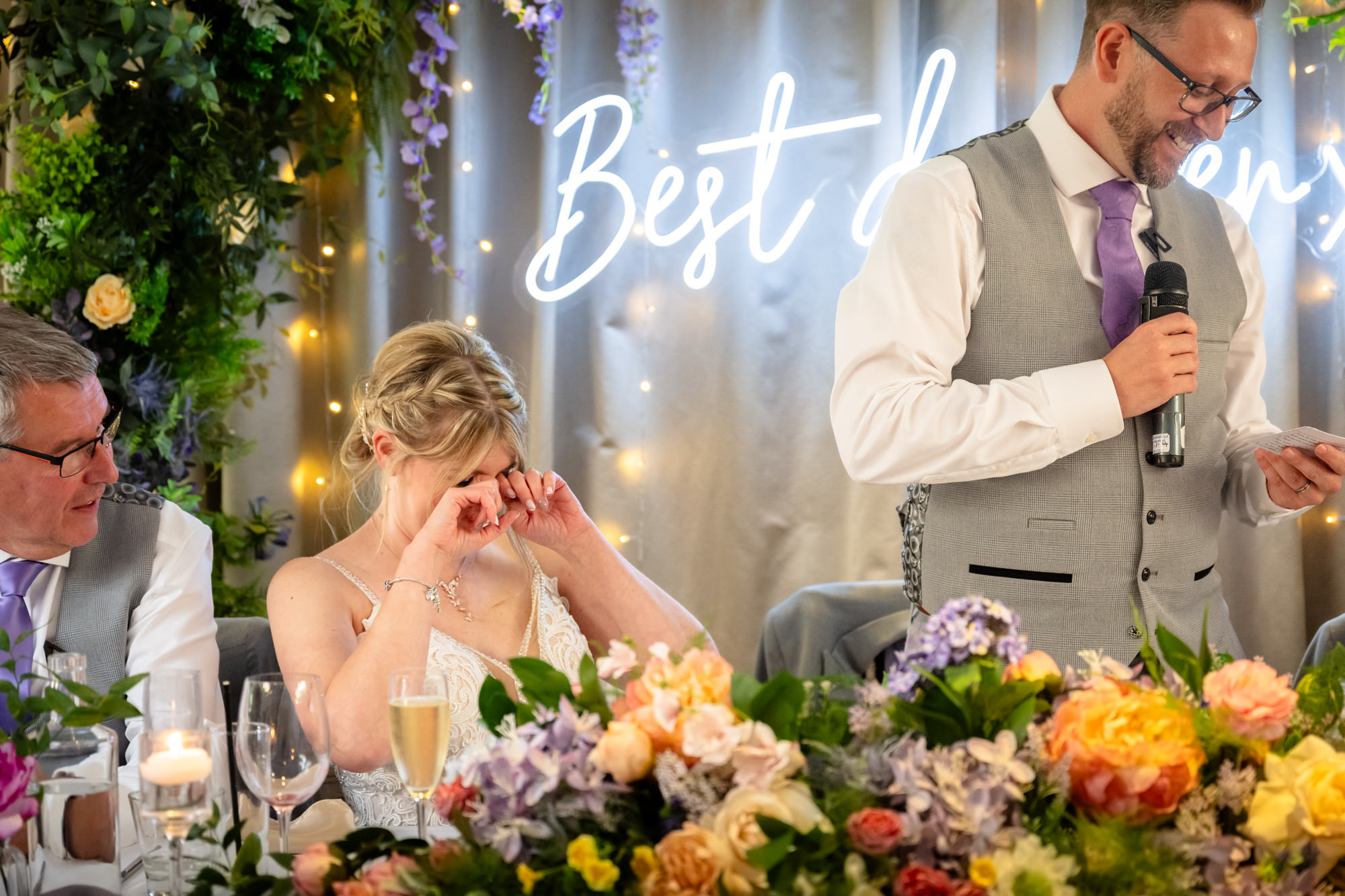 The groom speaks into a microphone during his speech at Moddershall Oaks, with a glowing neon sign in the background, visible through a floral arch.