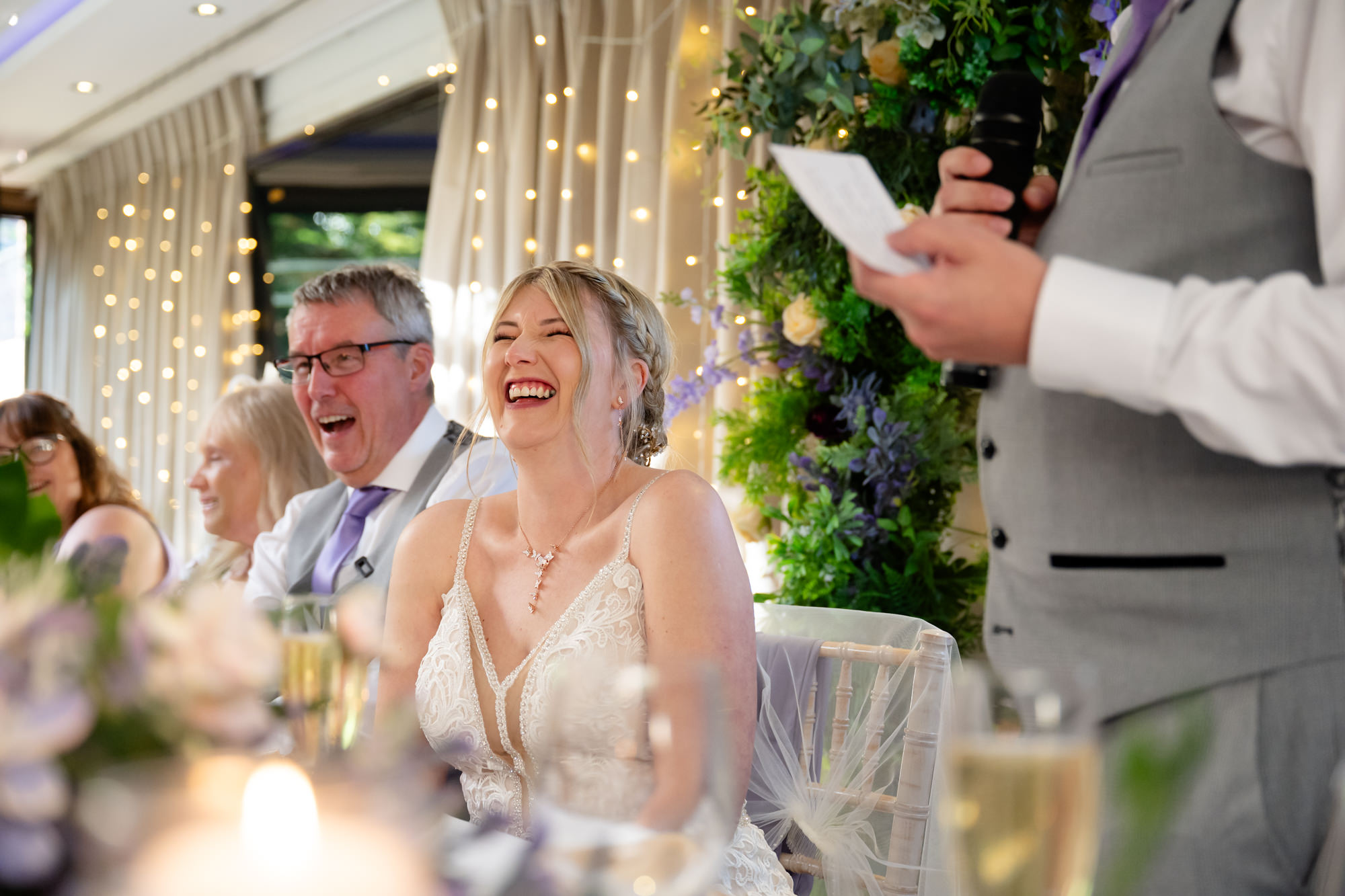 The bride and groom are laughing heartily at the head table at Moddershall Oaks, with guests blurred in the background, during the speeches.