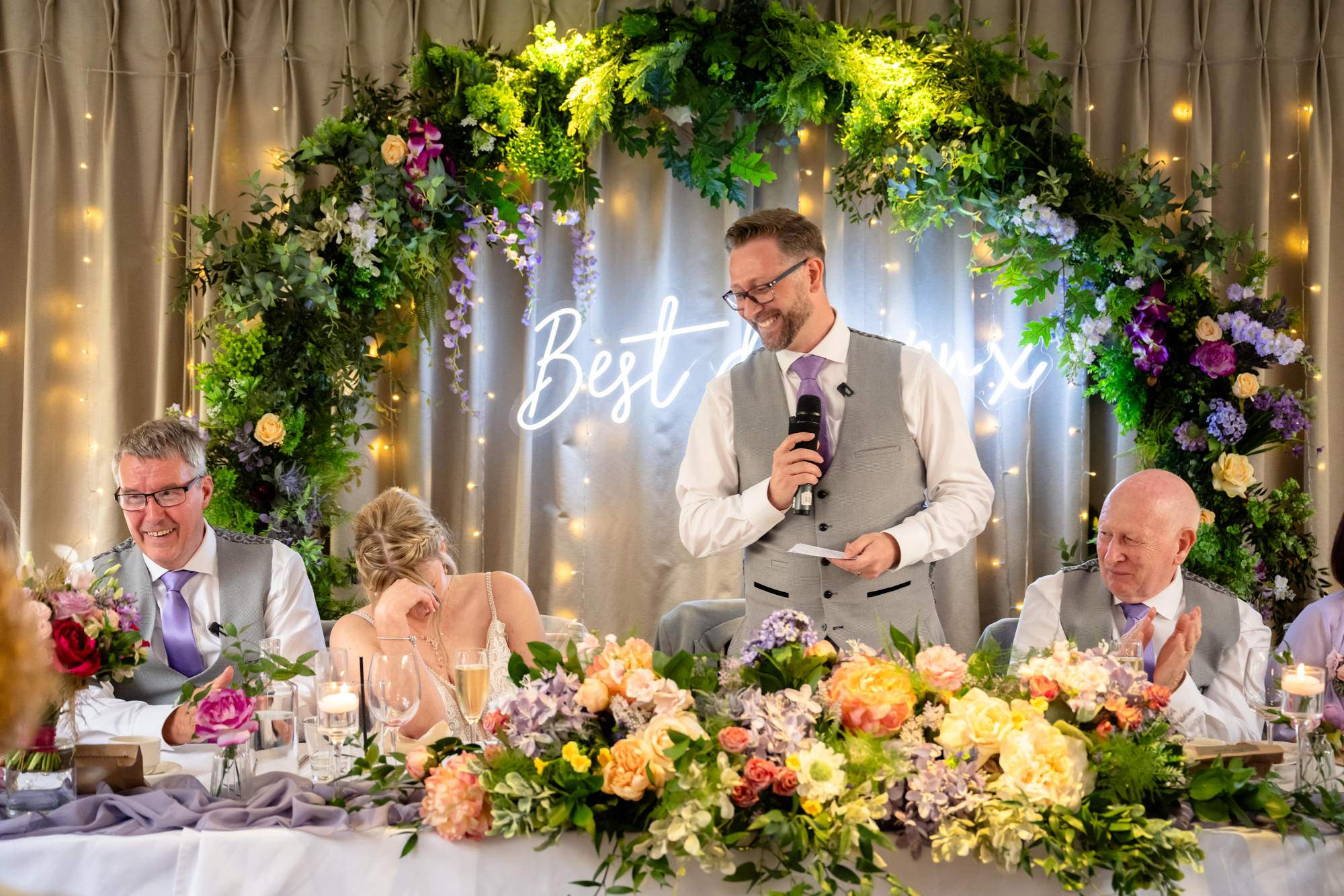 The groom speaks confidently into a microphone at the head table at Moddershall Oaks, with a vibrant floral arch behind him and guests seated, during his speech.