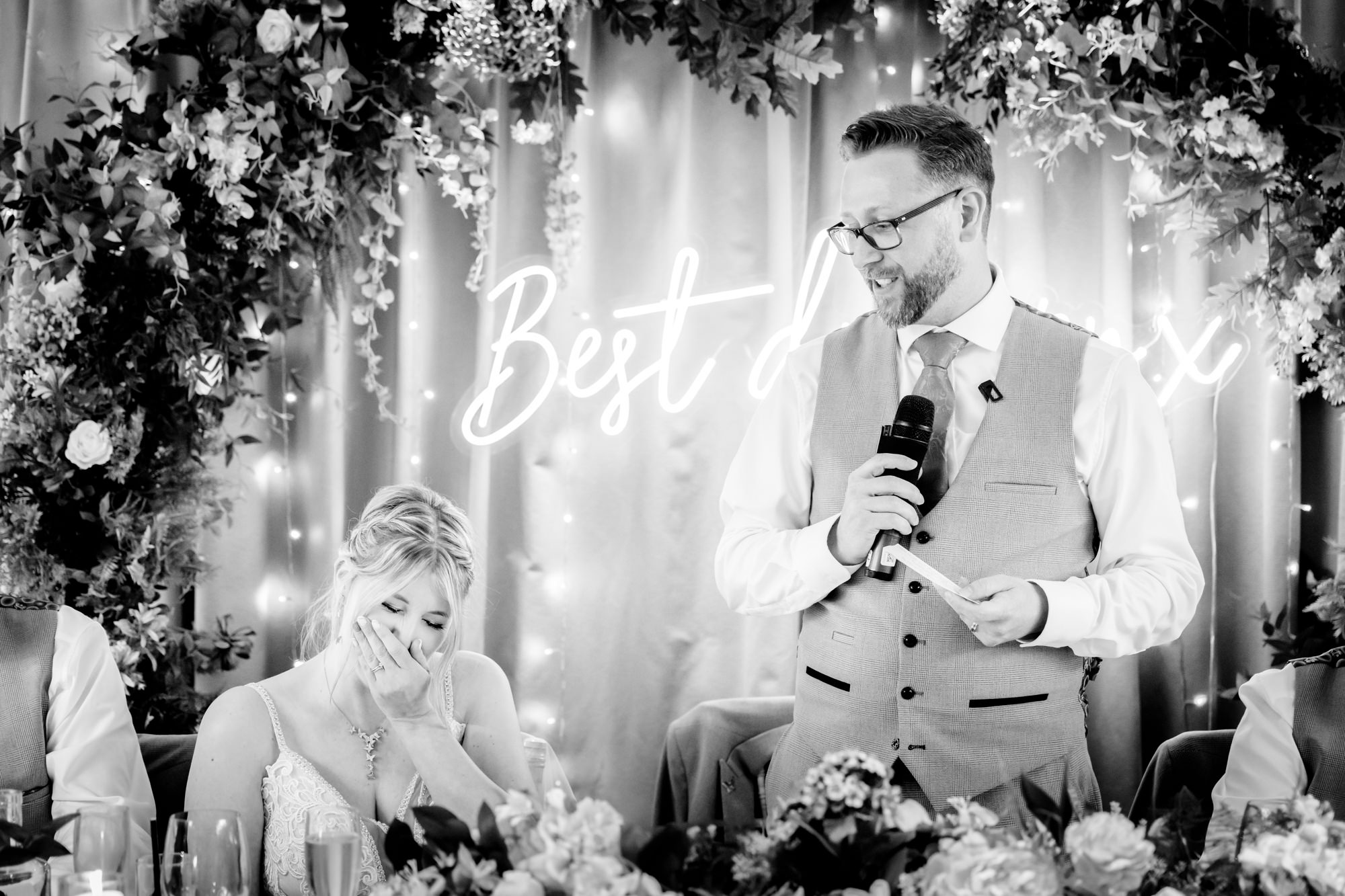 A black and white image of the groom speaking at the head table at Moddershall Oaks, looking out at the guests during his speech.