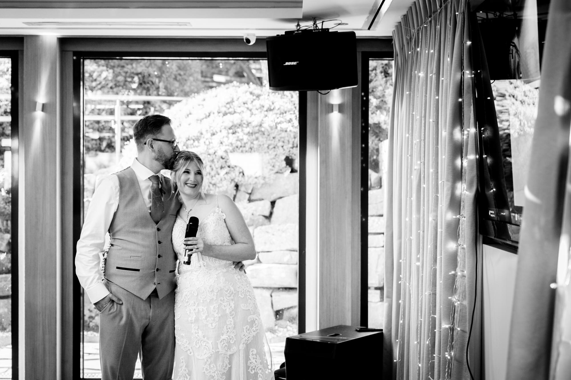 A black and white image of the bride and groom standing together, holding a microphone and smiling, watching the bride's ice skating routine at Moddershall Oaks.