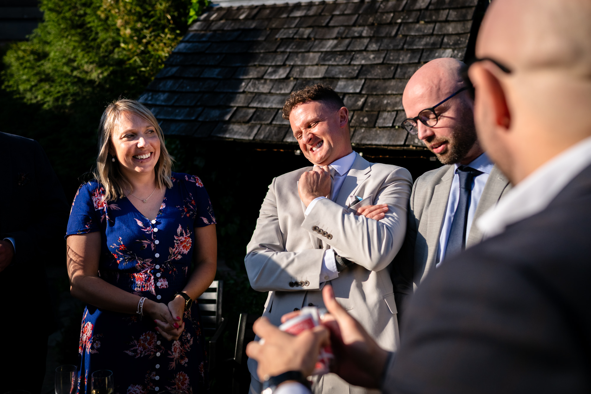 Three guests, including the groom in his grey suit, are seen from the chest up, laughing together outdoors in the sun, entertained by close up magic at Moddershall Oaks.