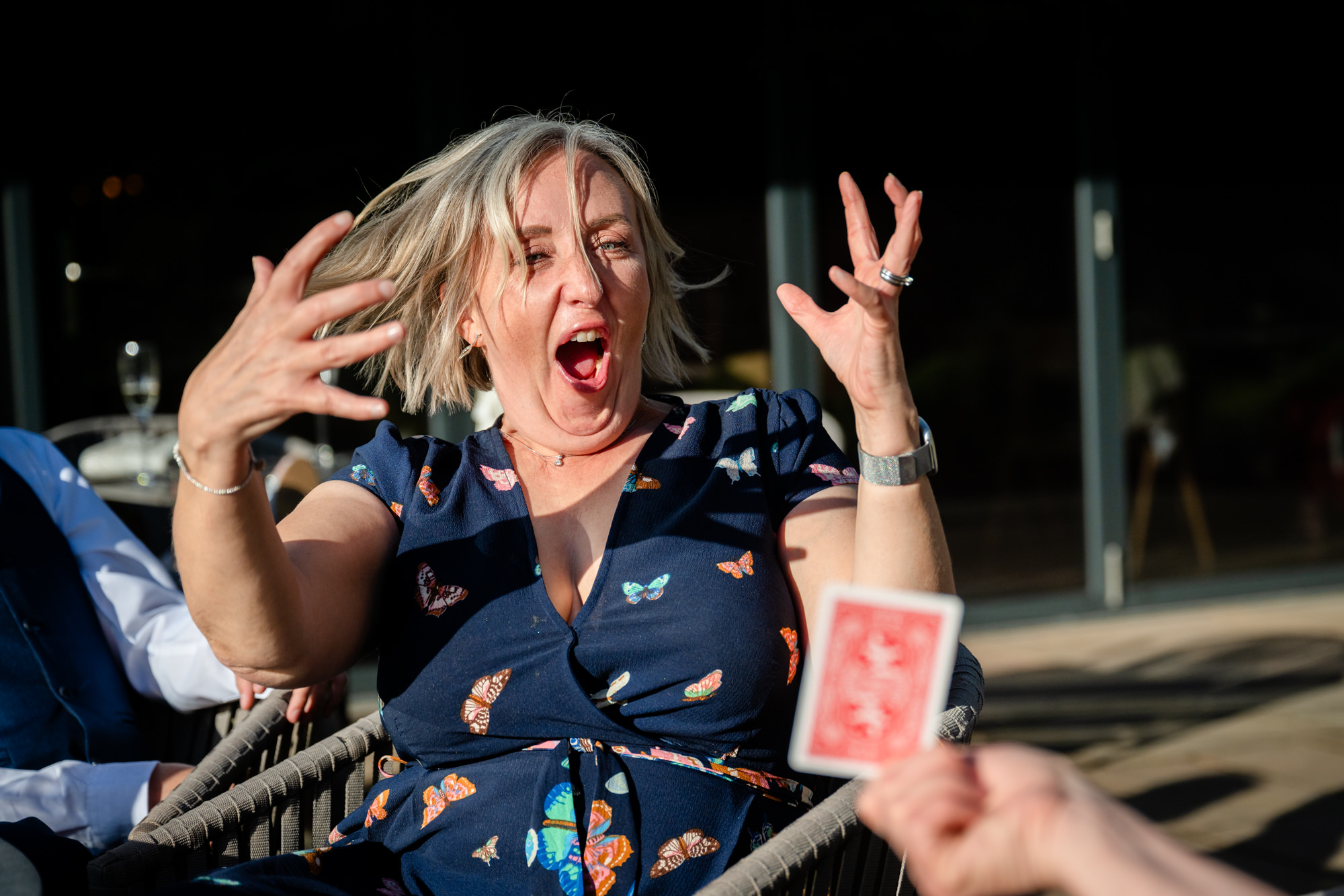 A woman with vibrant pink nails throws her hands up and shouts with excitement, captured in a candid, high-energy moment during close up magic entertainment at Moddershall Oaks.