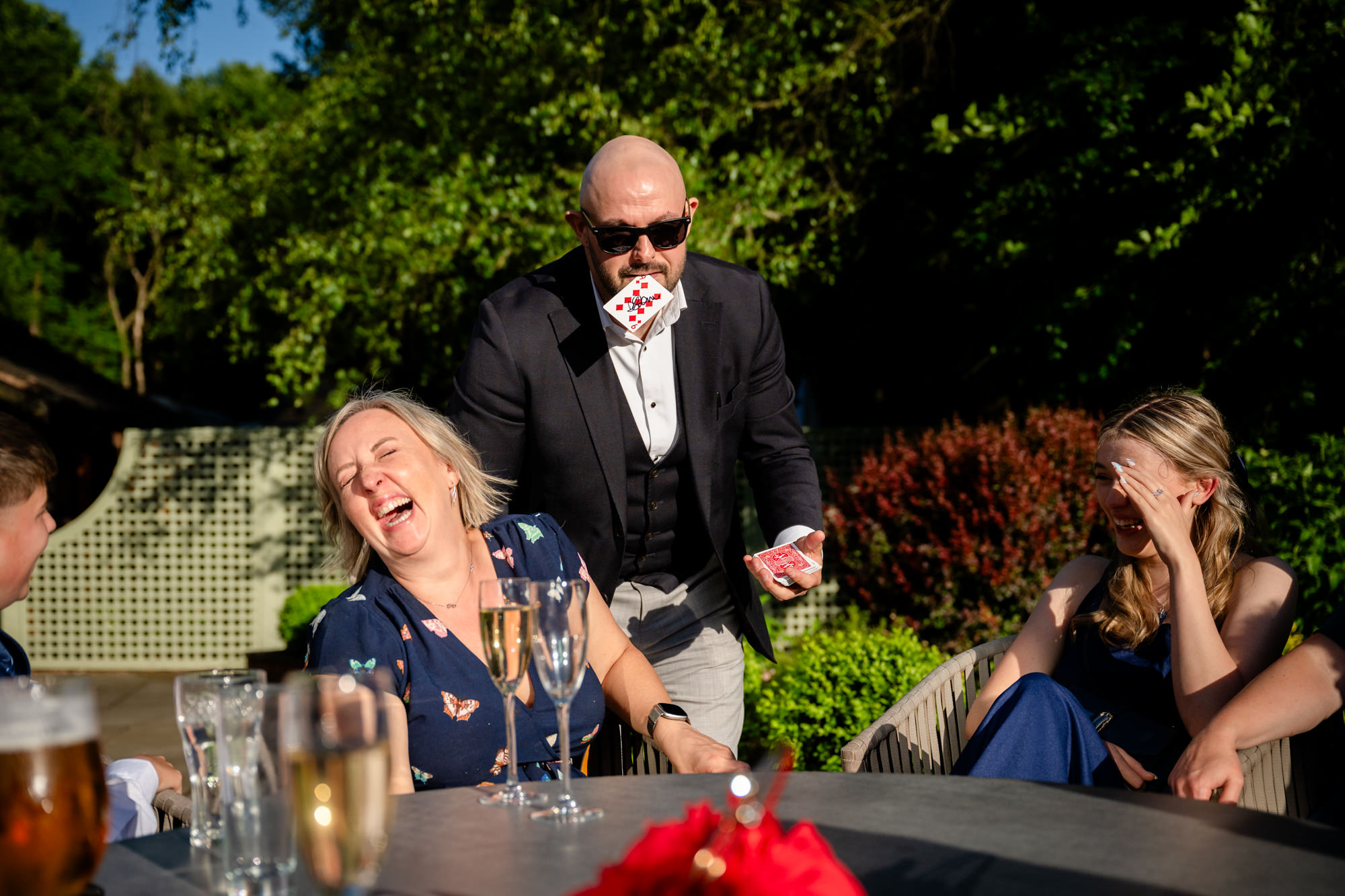 A man in a suit, wearing sunglasses and a card in his mouth, performs a magic trick for guests outdoors at Moddershall Oaks, with trees in the background, during close up magic entertainment.