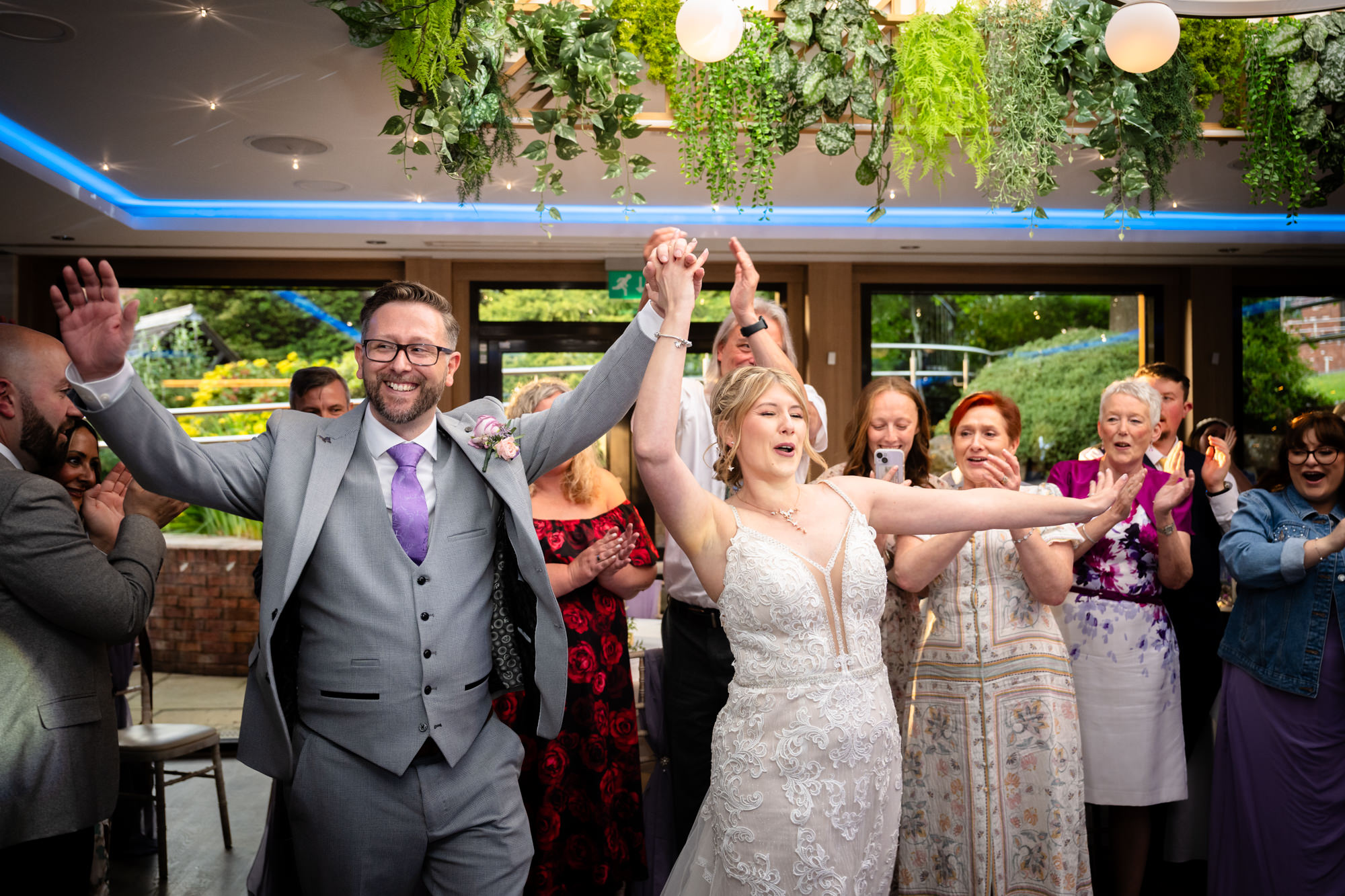 The bride and groom, along with guests, dance and cheer with their arms raised, celebrating on the dance floor during the first dance at Moddershall Oaks.