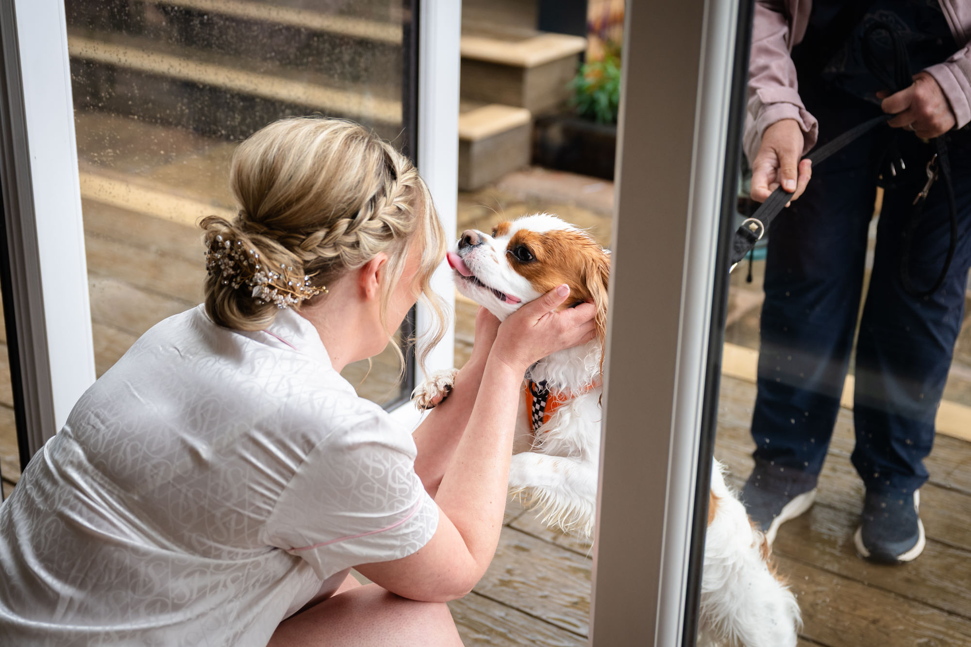 A bride in a white robe kneeling to pet a small, fluffy dog through a glass door, both showing affection, during bridal prep in the prep pod at Moddershall Oaks.