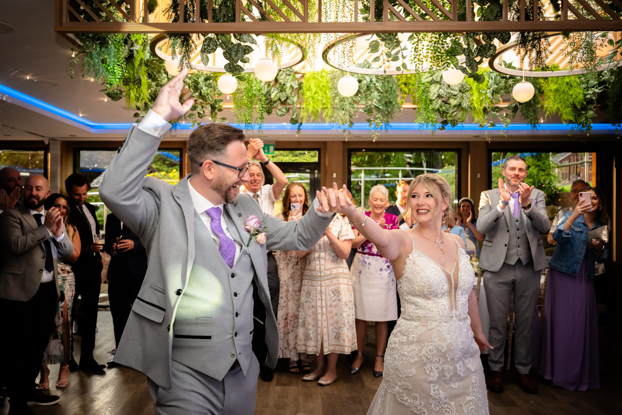 The bride and groom dance and smile with their arms raised, surrounded by cheering guests on the dance floor, during their first dance at Moddershall Oaks.