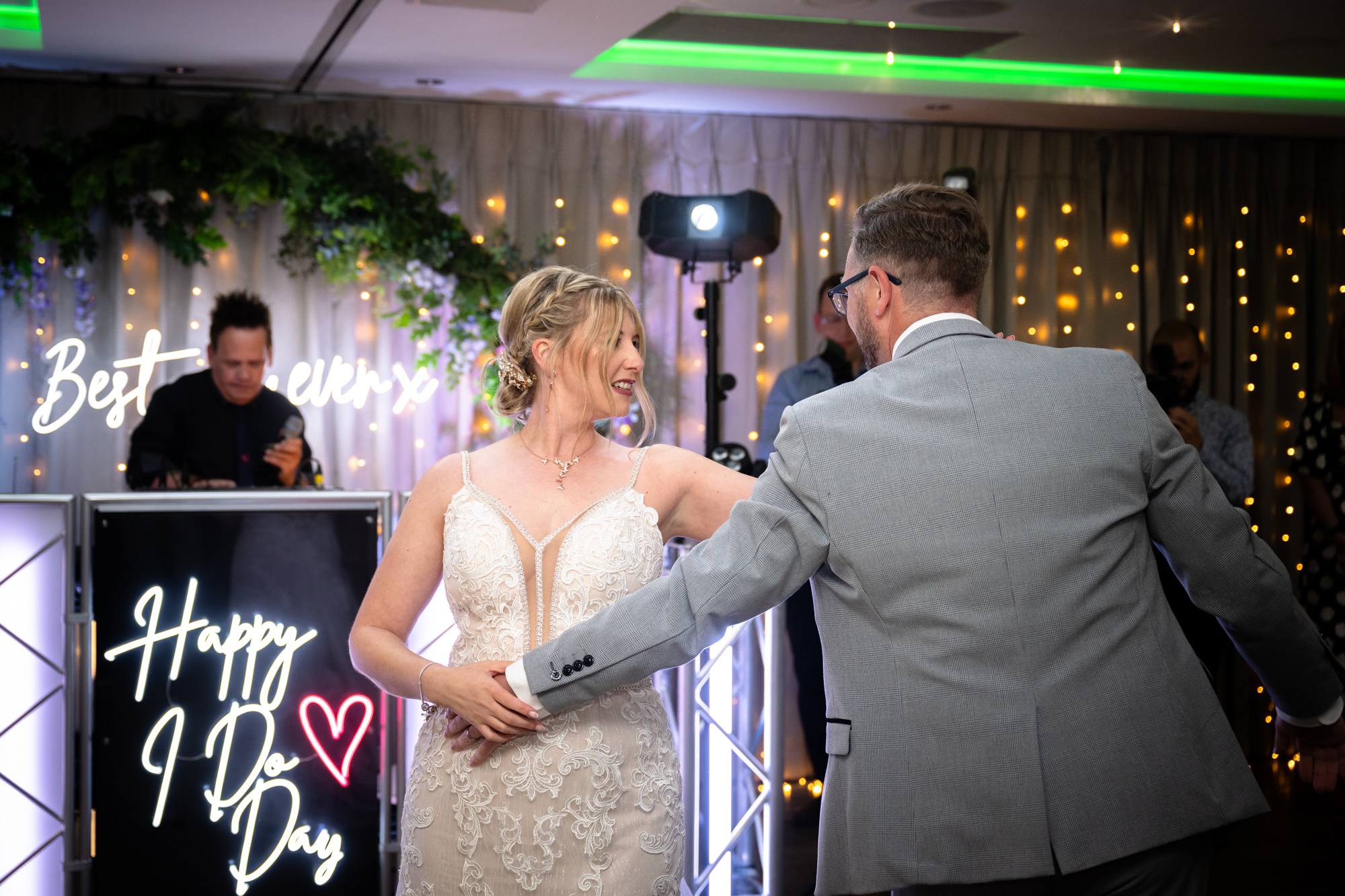 The groom carries the bride in his arms while dancing, with colorful lights and guests in the background, during the first dance at Moddershall Oaks.