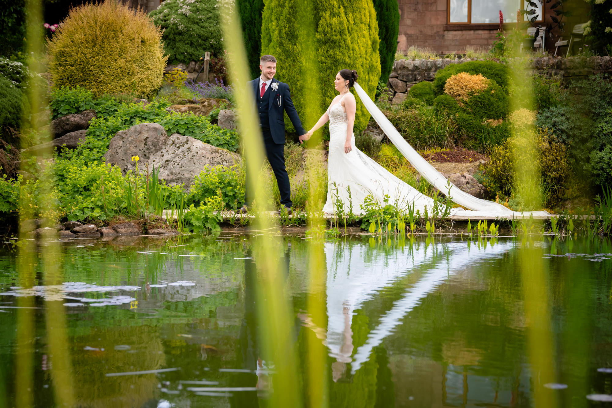 A portrait photo of the bride and groom standing near a pond at Heaton House Farm, their reflections visible in the water.