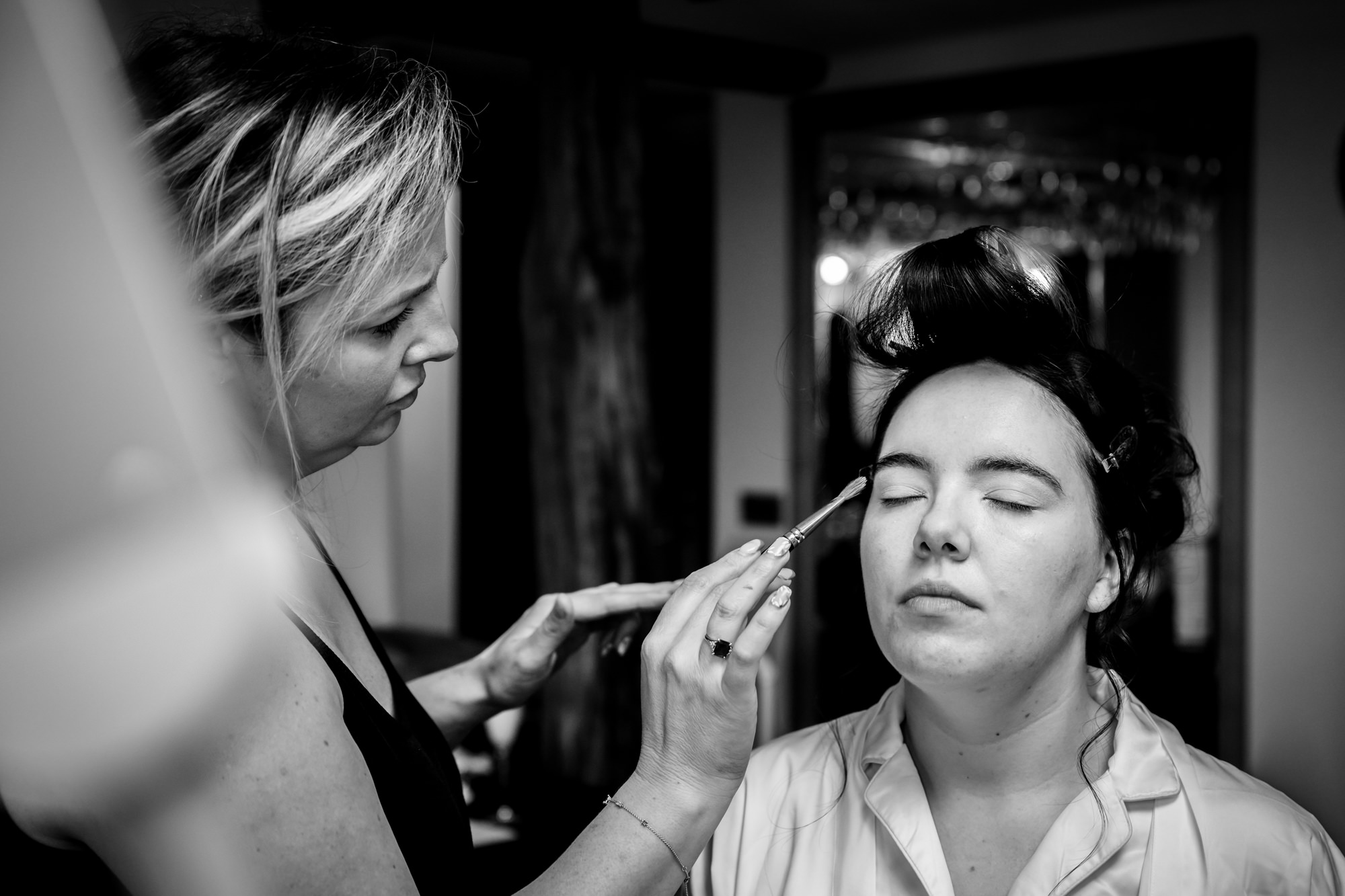 A close-up, black and white photo shows a woman applying eyeshadow to the bride in the Pamper room at Heaton House Farm.