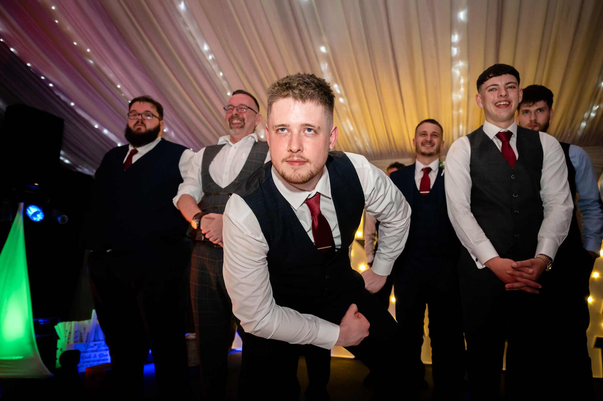 A group of men in vests and ties are dancing together at the Heaton House Farm wedding party.