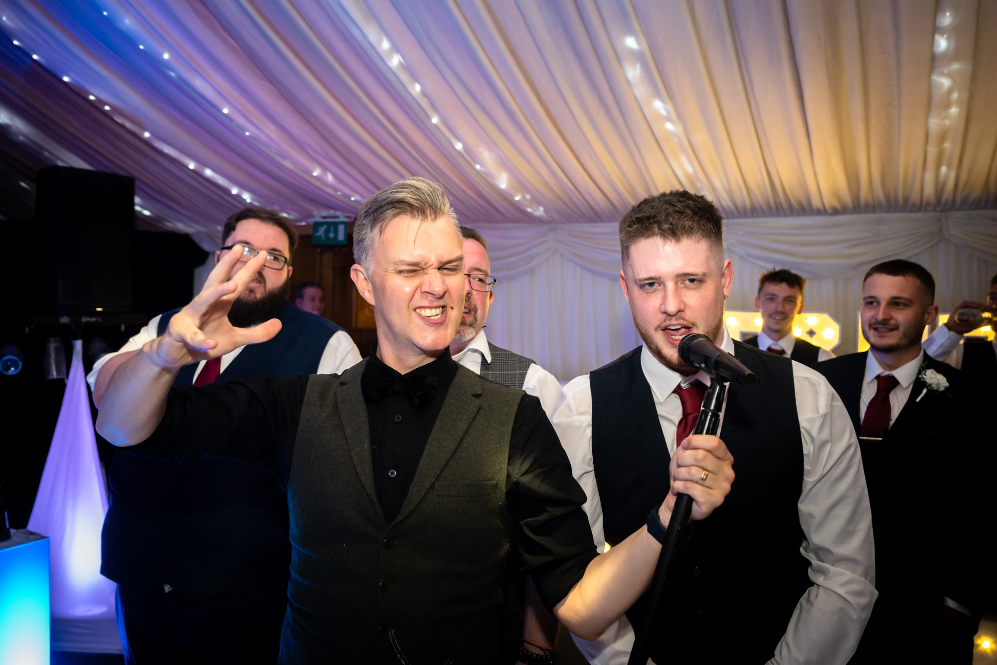 Two men are laughing together while a third man is singing into a microphone in the background, a moment from the wedding party at Heaton House Farm.