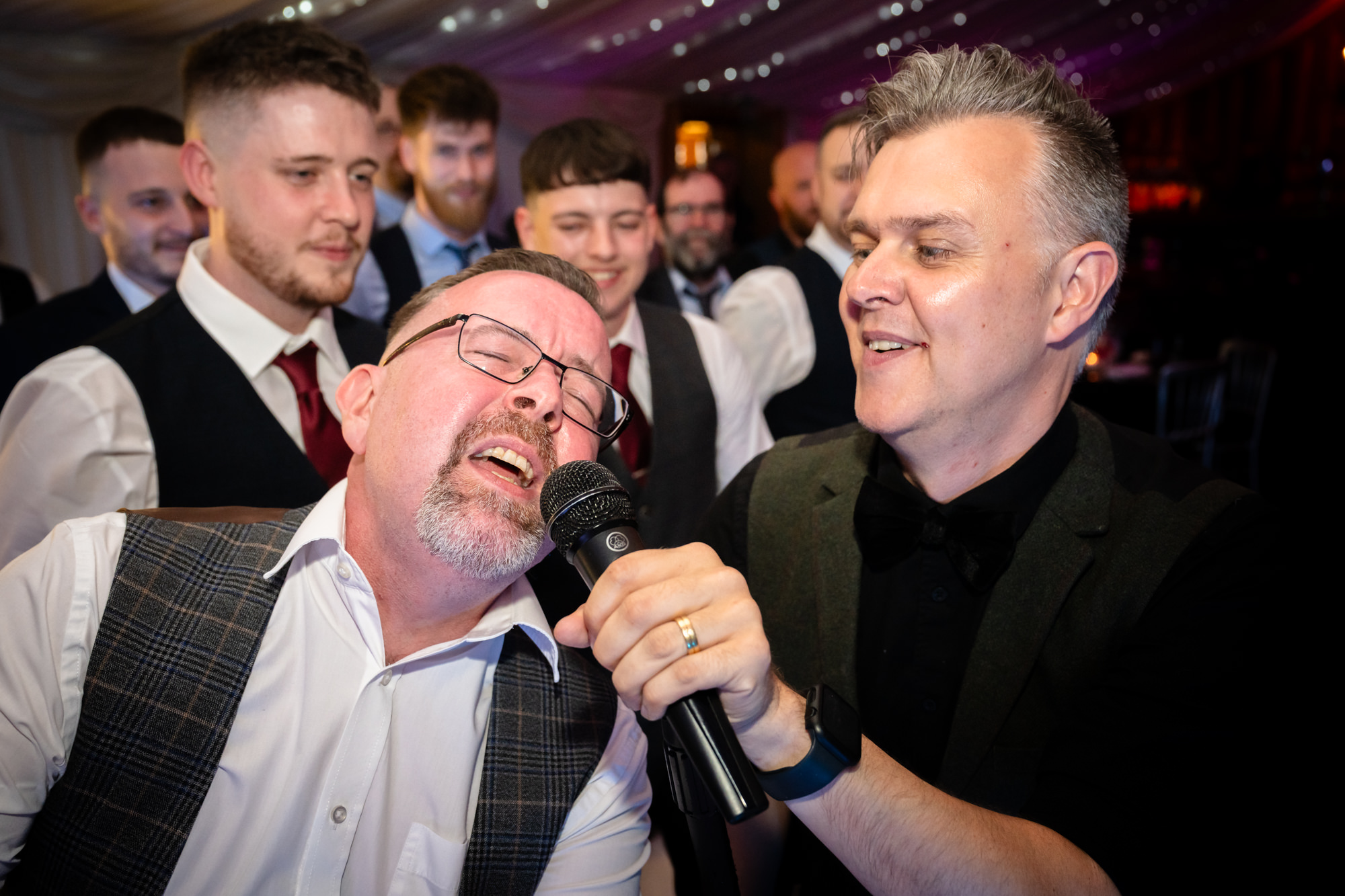 Two men smile and sing into a microphone together at the Heaton House Farm wedding party.