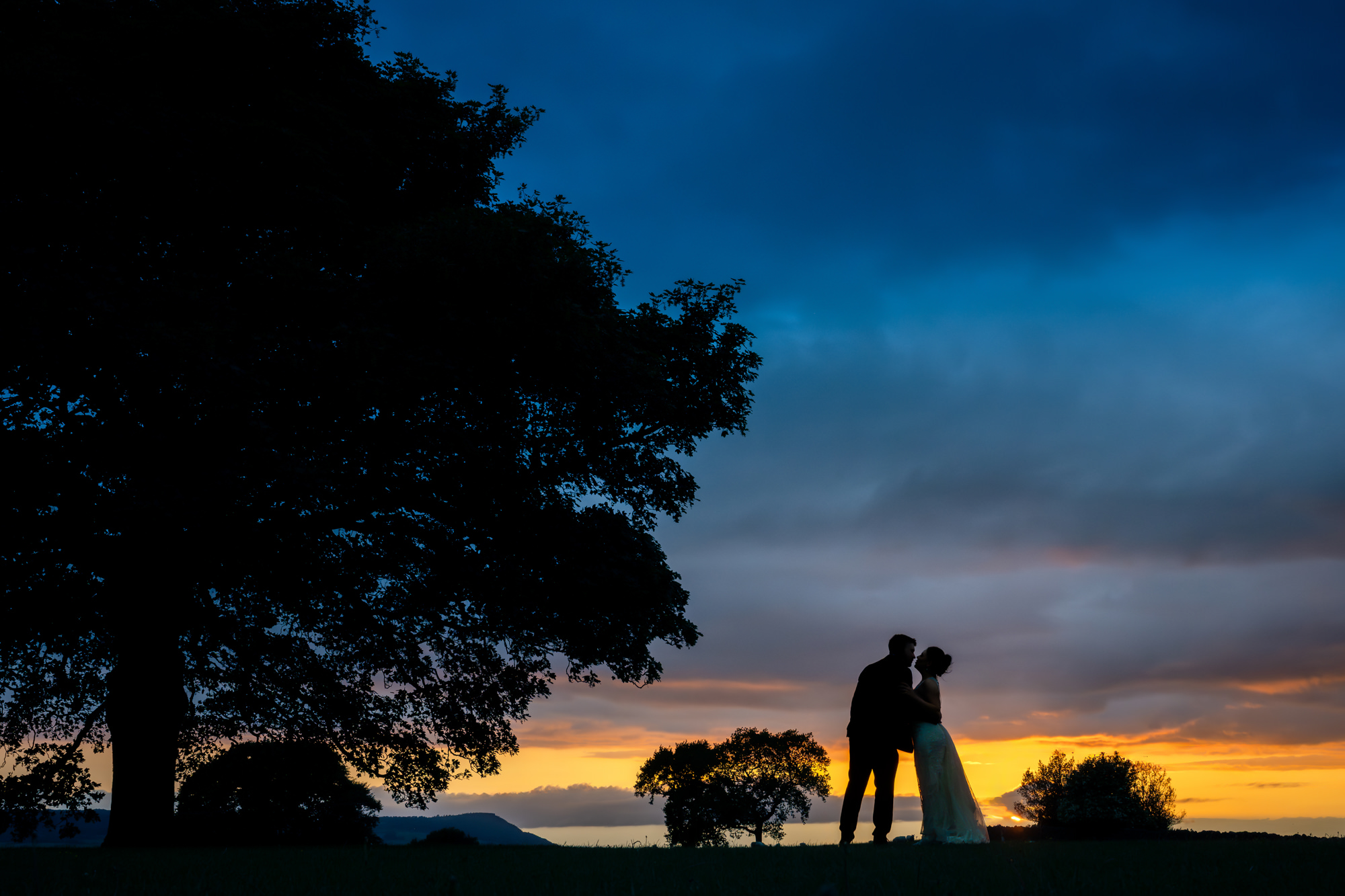 A beautiful silhouette of the bride and groom standing together under a tree against a vibrant sunset sky at Heaton House Farm.