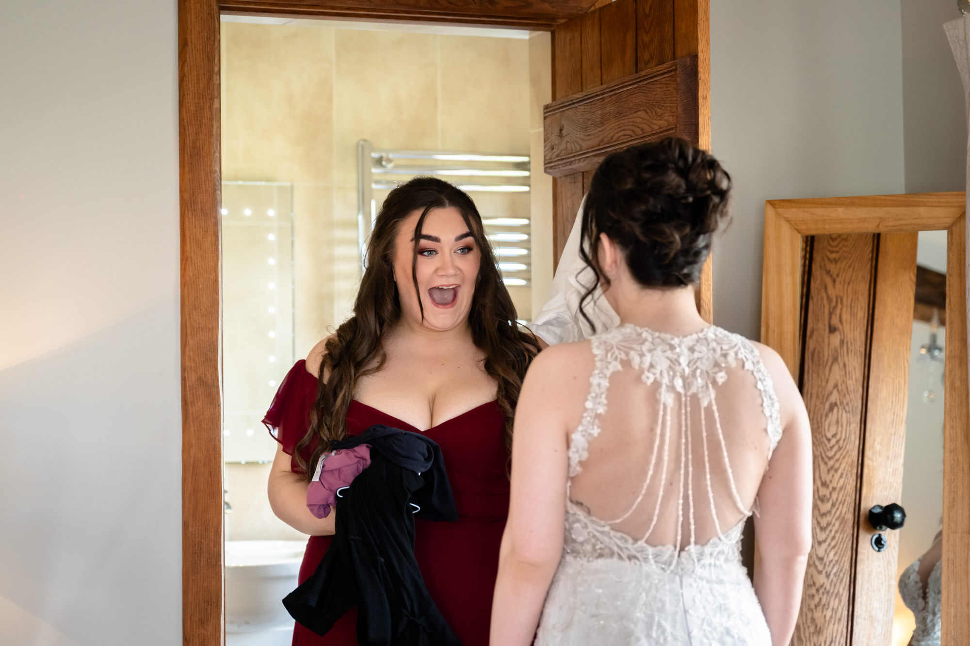 A bridesmaid in a red dress looks surprised and excited while looking at the bride, whose back is to the camera. The photo was taken at Heaton House Farm.