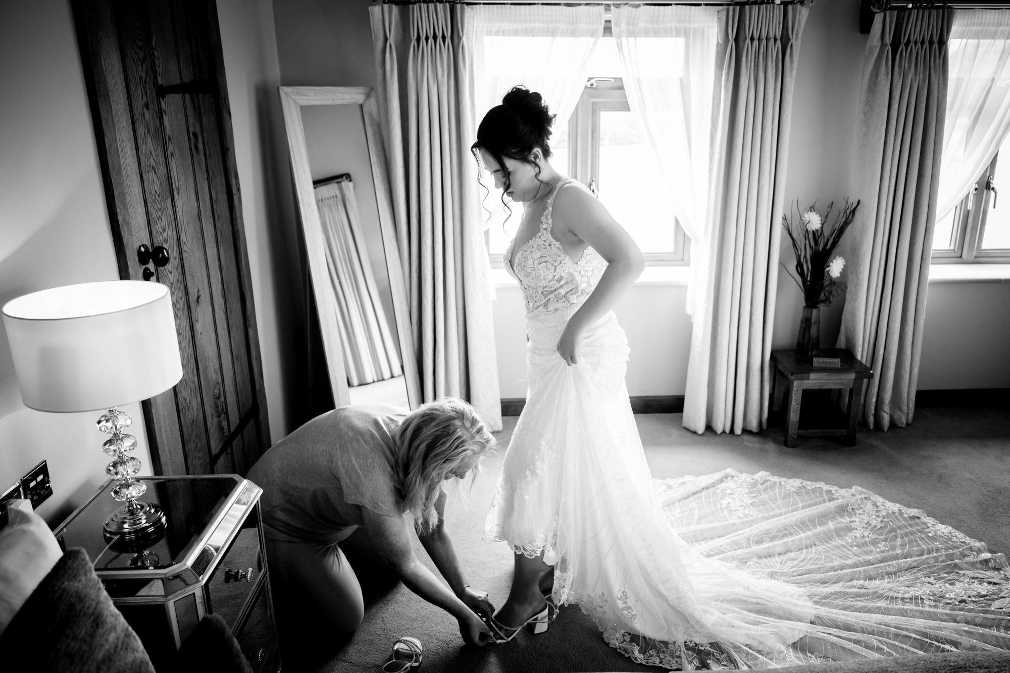 A black and white photo shows a woman kneeling to help the bride put on her shoes, as the bride stands in her dress at Heaton House Farm.