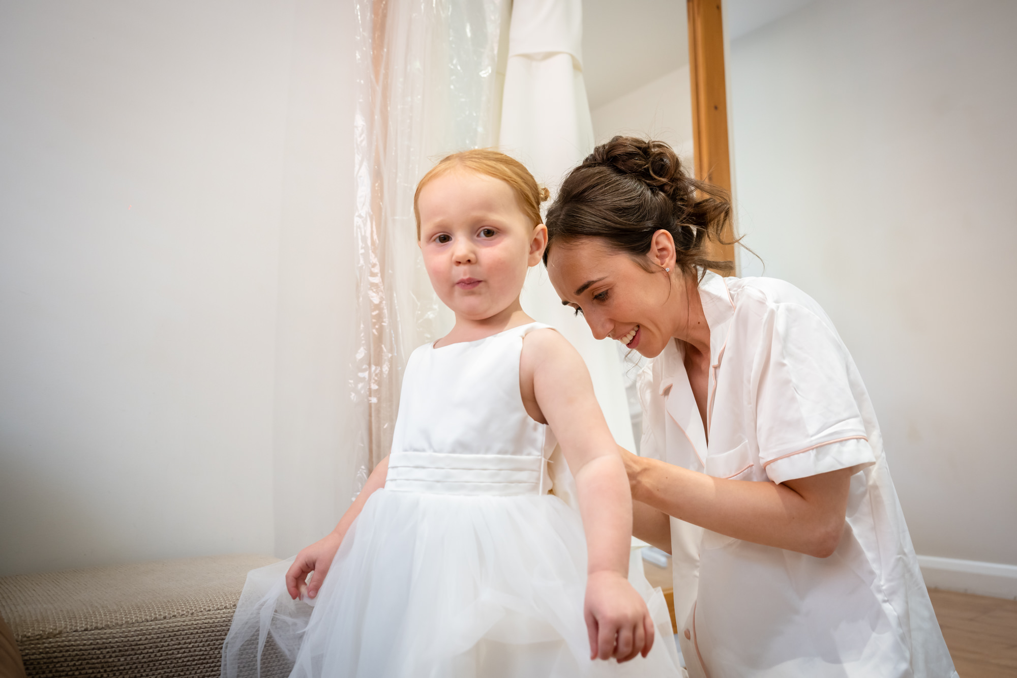 The bride helps a young girl in a white dress get ready in the dressing room at Hanbury Barns.