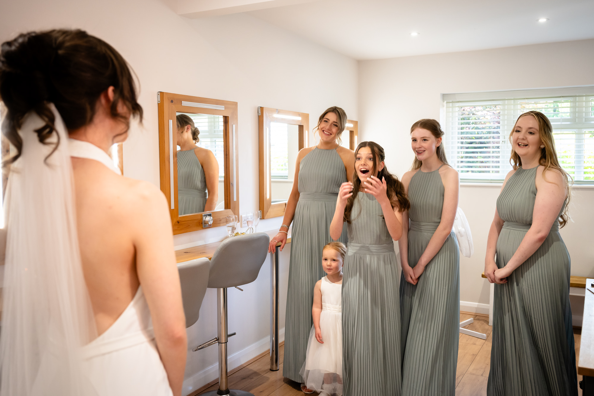 The bride, whose back is to the camera, is seen with a group of bridesmaids in gray dresses who are smiling and looking at her, in the dressing room at Hanbury Barns.